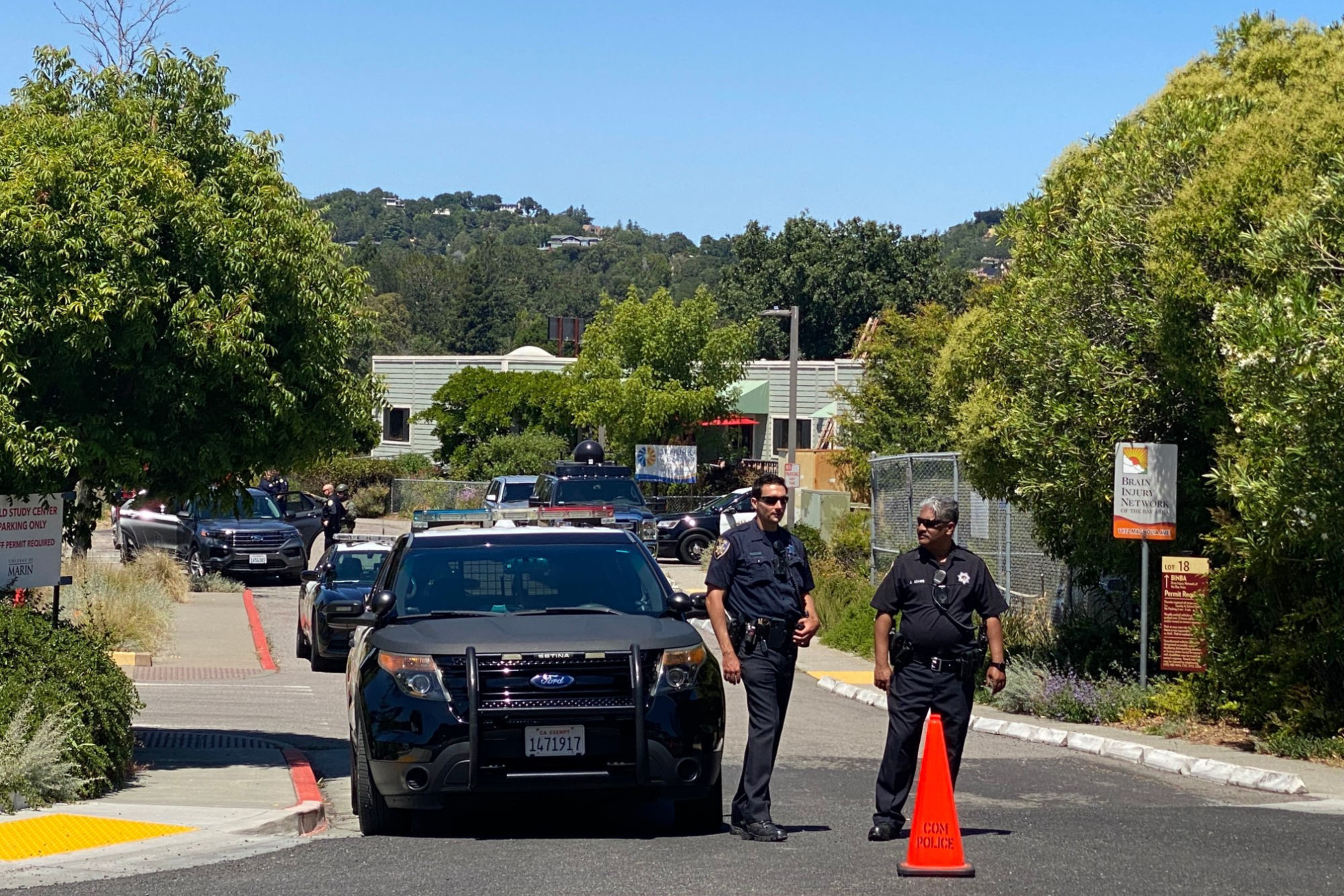 Two police officers stand beside a black police SUV blocking a road with trees and buildings in the background on a sunny day.