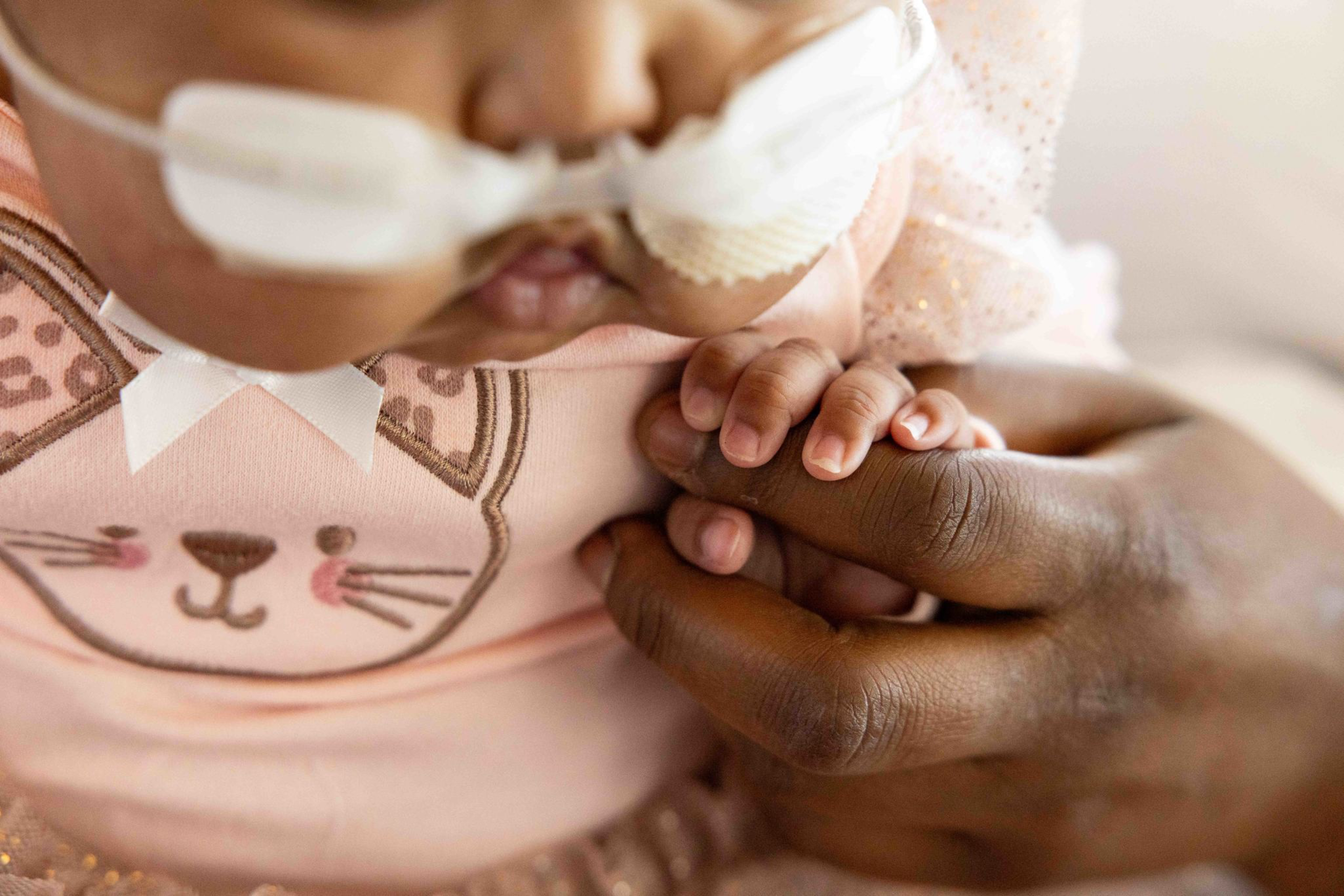 A baby wearing a pink outfit with a cat face design has a nasal tube and gently holds an adult's finger.