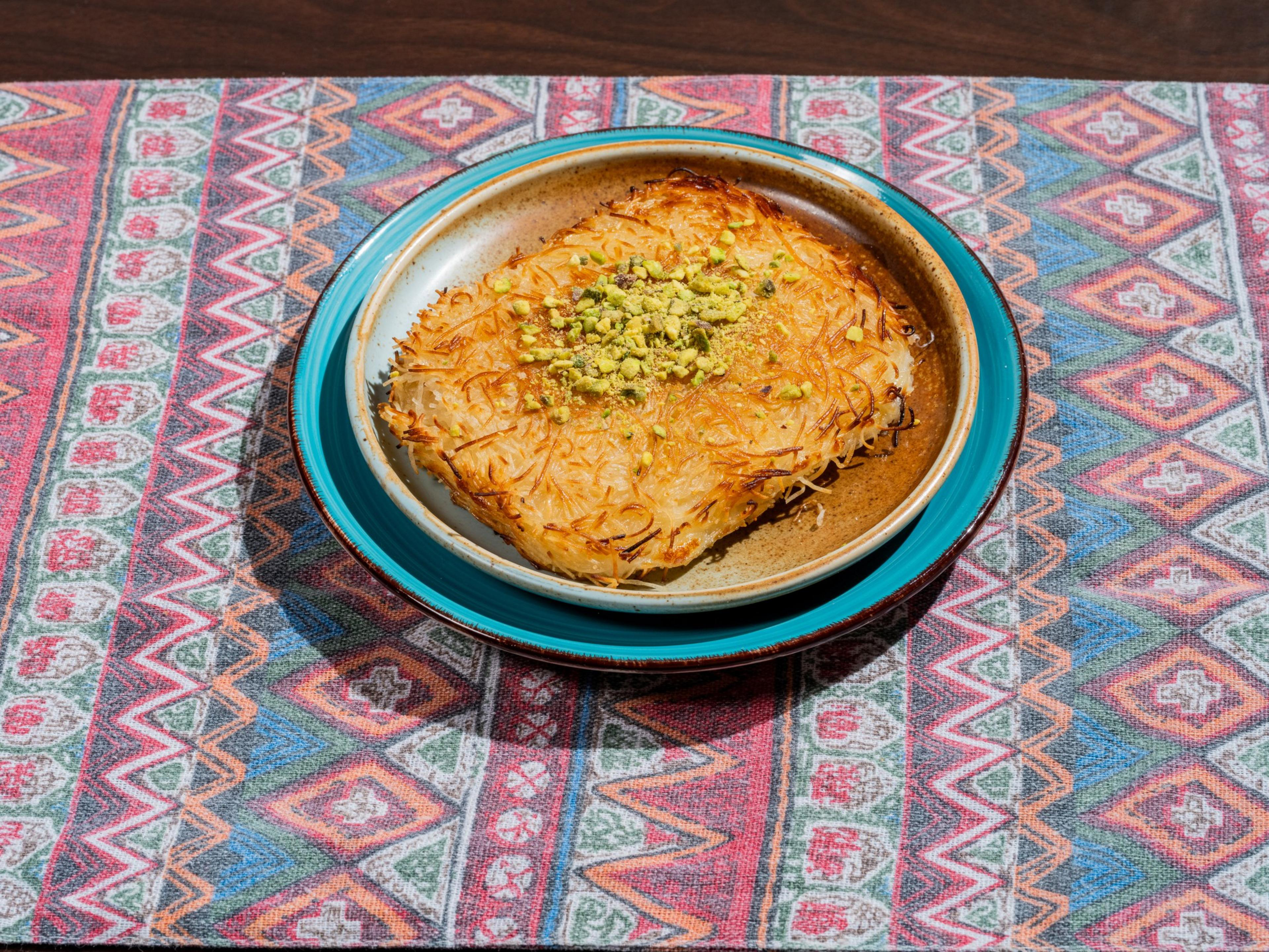 A baked dessert topped with crushed pistachios is on a blue-edged plate. The background is a colorful, geometric-patterned fabric.