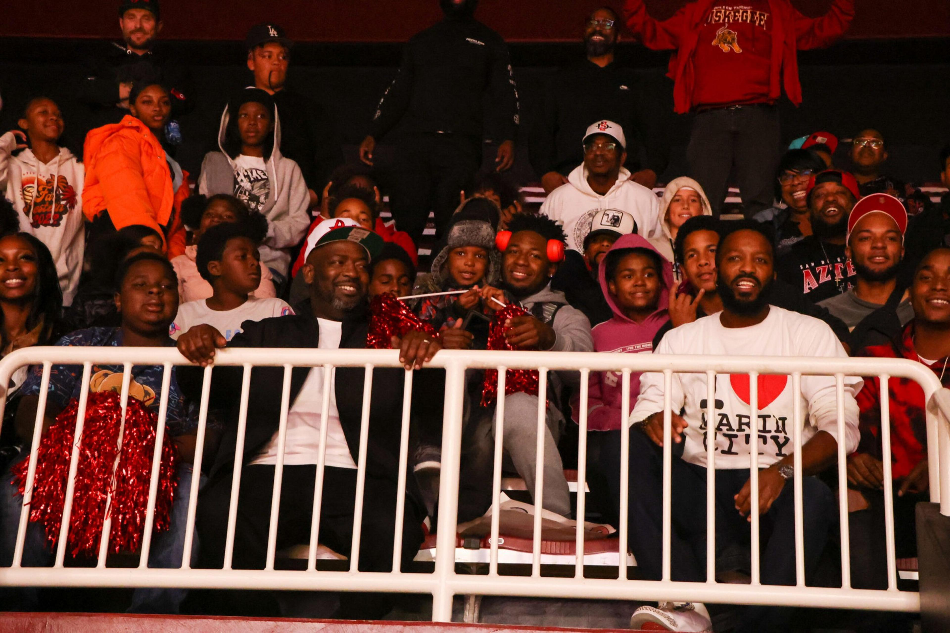 A diverse group of people, including children and adults, sit and stand behind a white railing, many smiling and some holding red pom-poms.