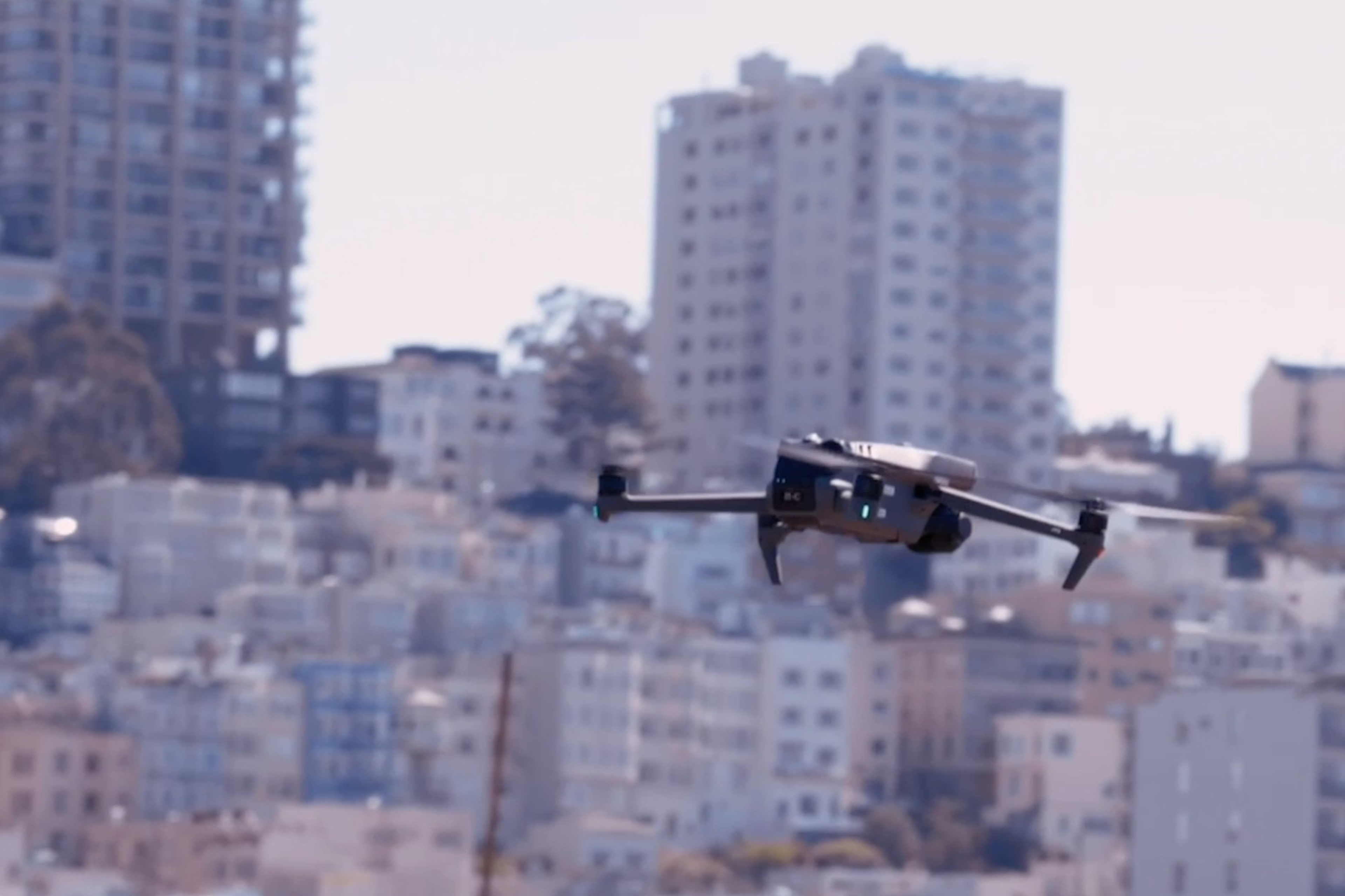 A drone is flying in front of a densely packed urban area with numerous tall buildings, possibly in San Francisco, as suggested by a visible police badge.