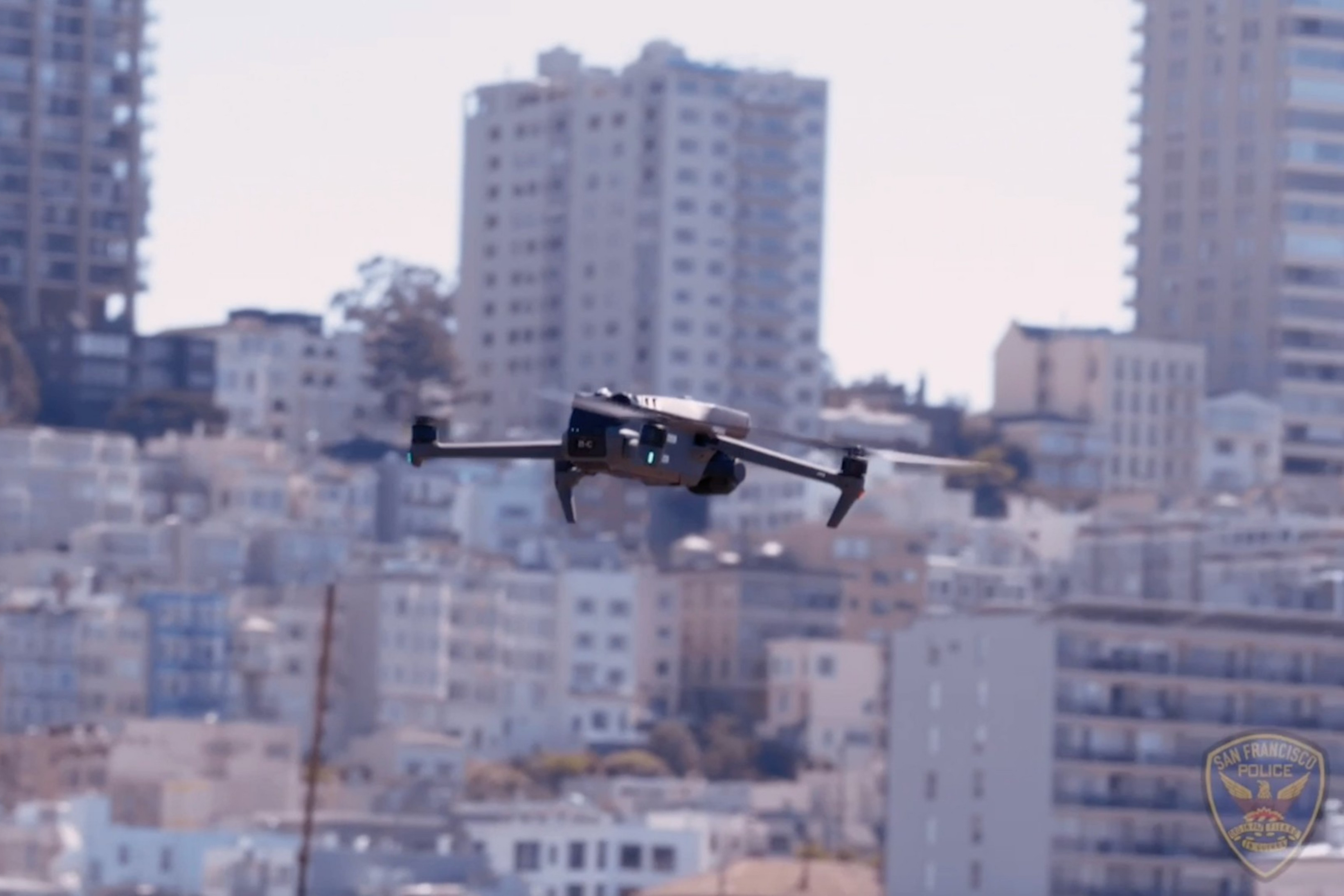 A drone is flying in front of a densely packed urban area with numerous tall buildings, possibly in San Francisco, as suggested by a visible police badge.