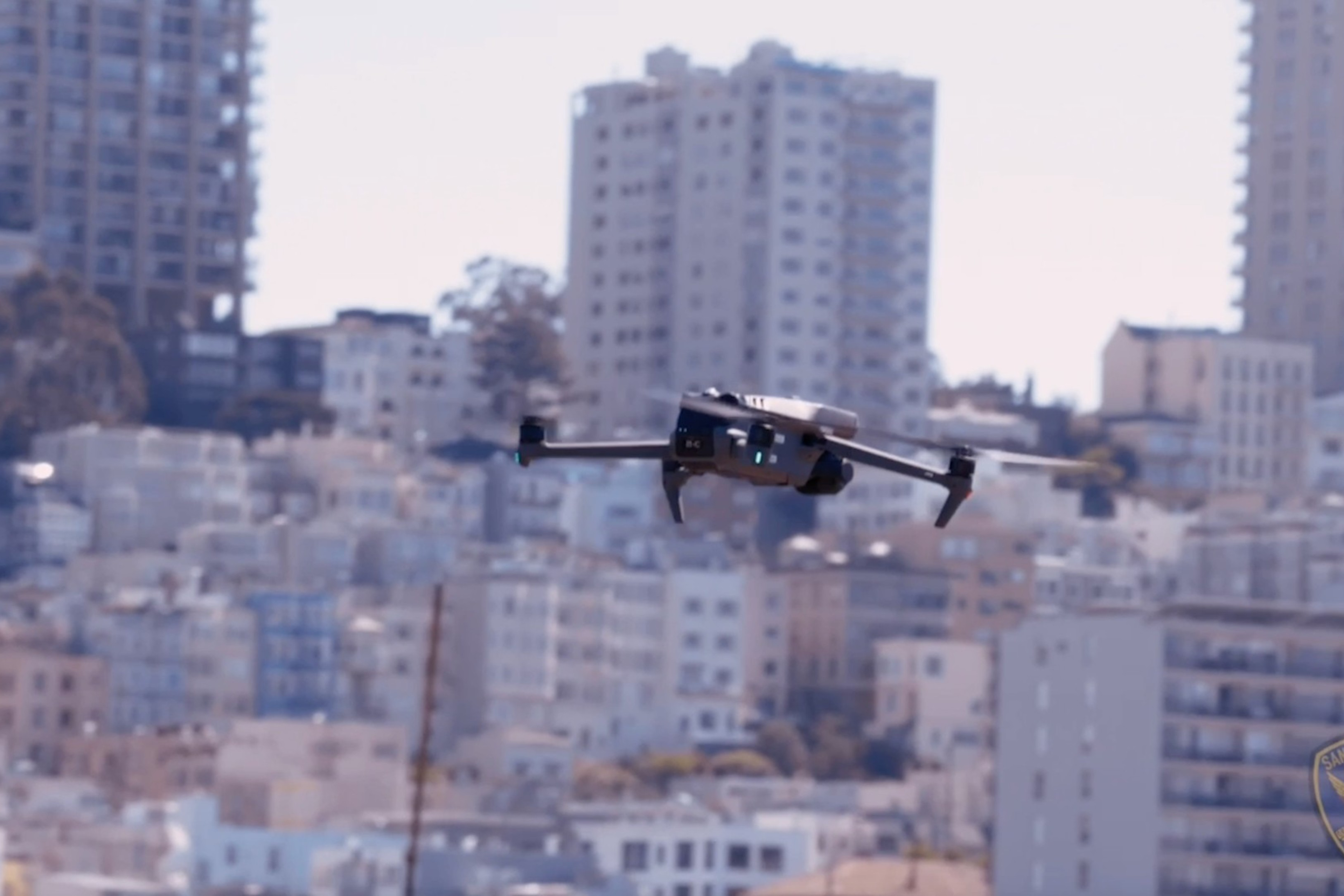 A drone is flying in front of a densely packed urban area with numerous tall buildings, possibly in San Francisco, as suggested by a visible police badge.