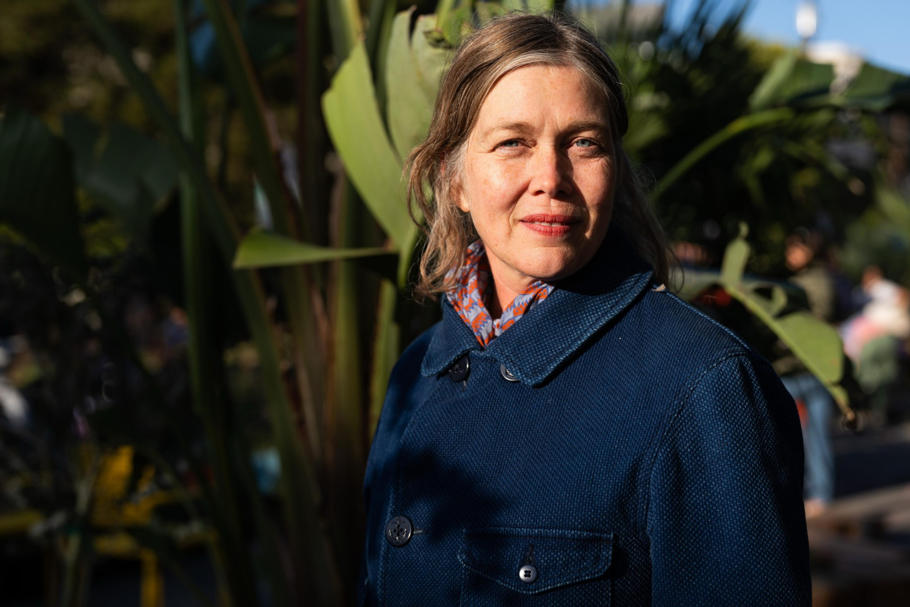 A woman in a blue coat stands outdoors in sunlight, surrounded by large green leaves. She has a calm expression and soft lighting highlights her face.