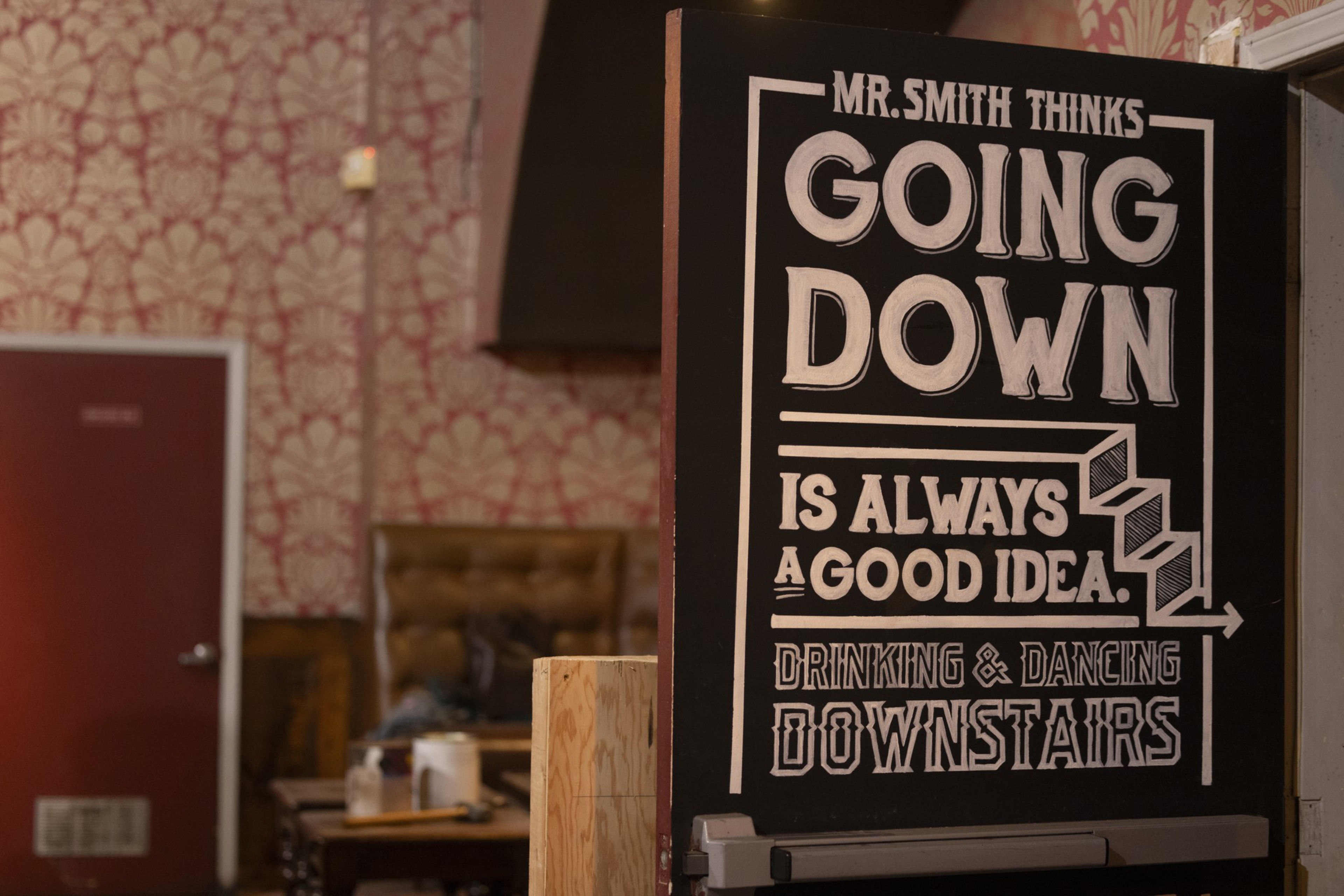 A black sign reads: &quot;Mr. Smith thinks going down is always a good idea. Drinking &amp; dancing downstairs.&quot; The background shows patterned wallpaper and a closed red door.