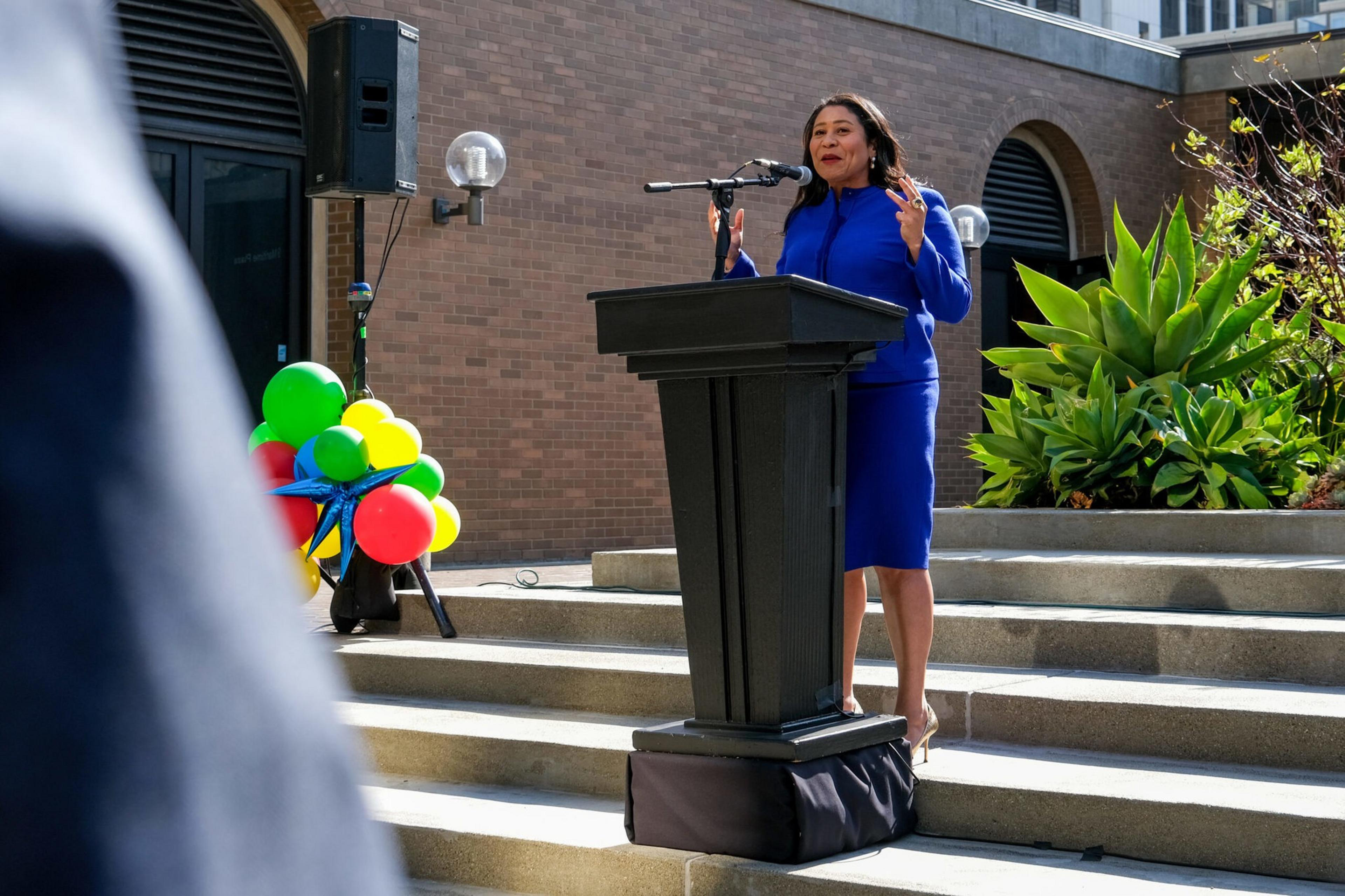 A woman in a blue dress speaks at a podium on outdoor steps, with colorful balloons and greenery in the background.