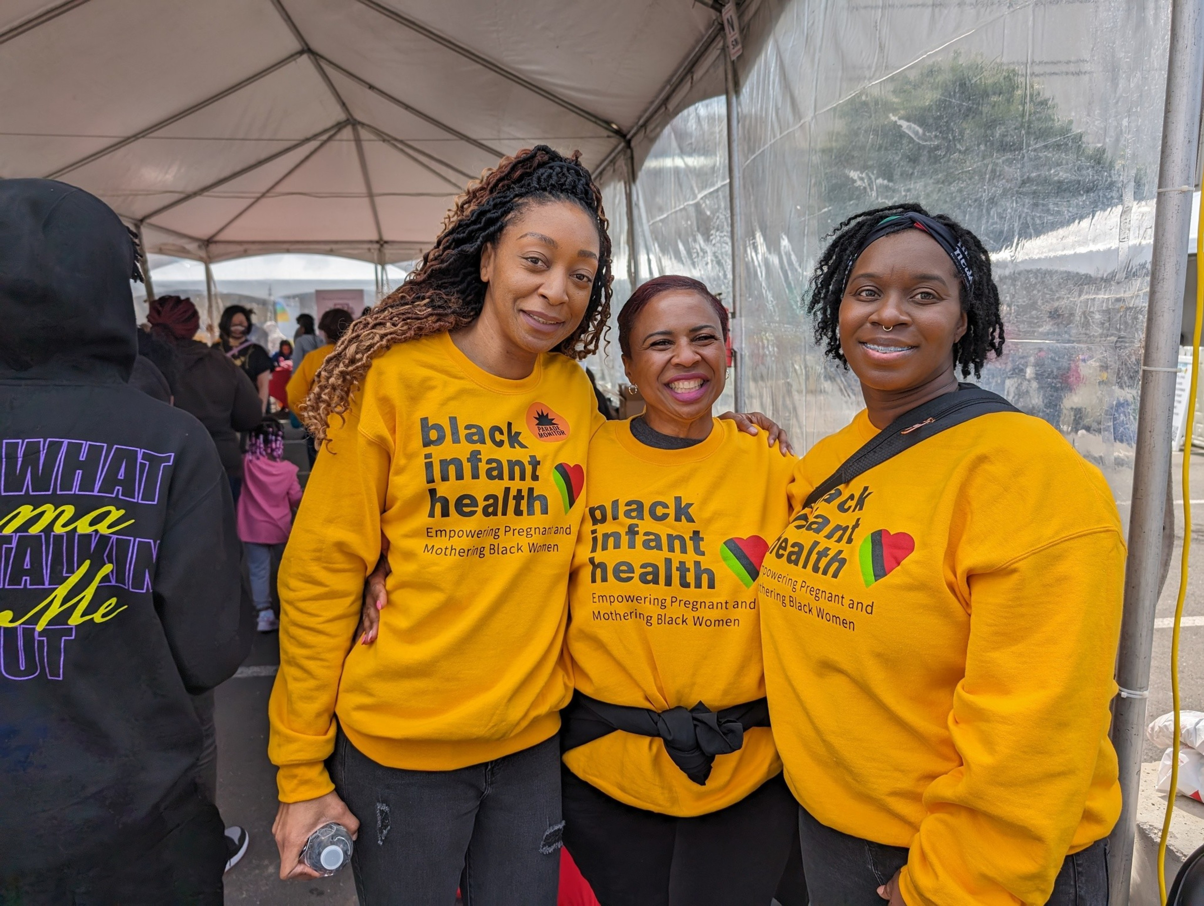 Three women in bright yellow sweatshirts stand at an outdoor festival booth.