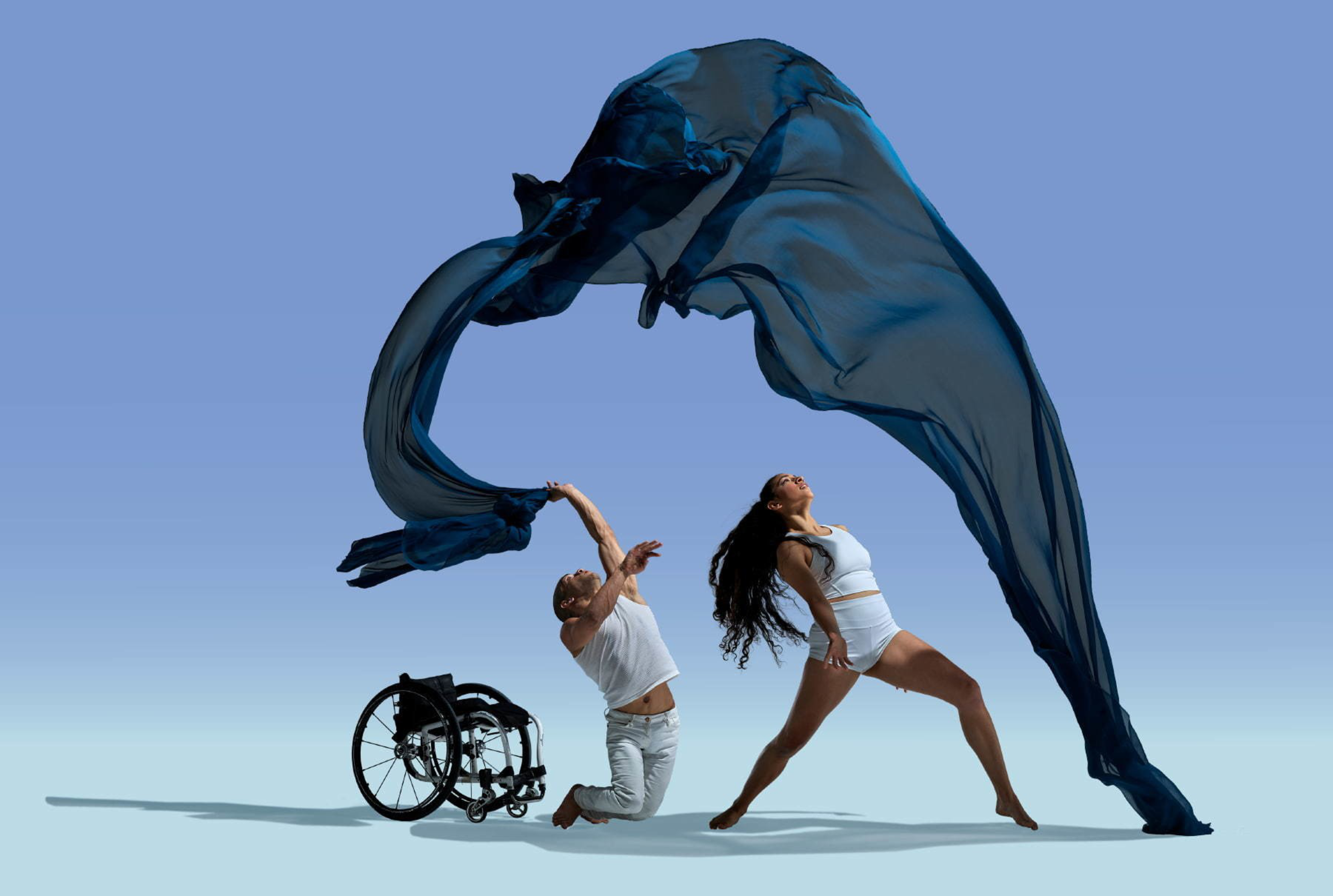 A man in a wheelchair is kneeling and throwing a flowing blue fabric into the air while a woman in white dancewear strikes a dynamic pose beside him.