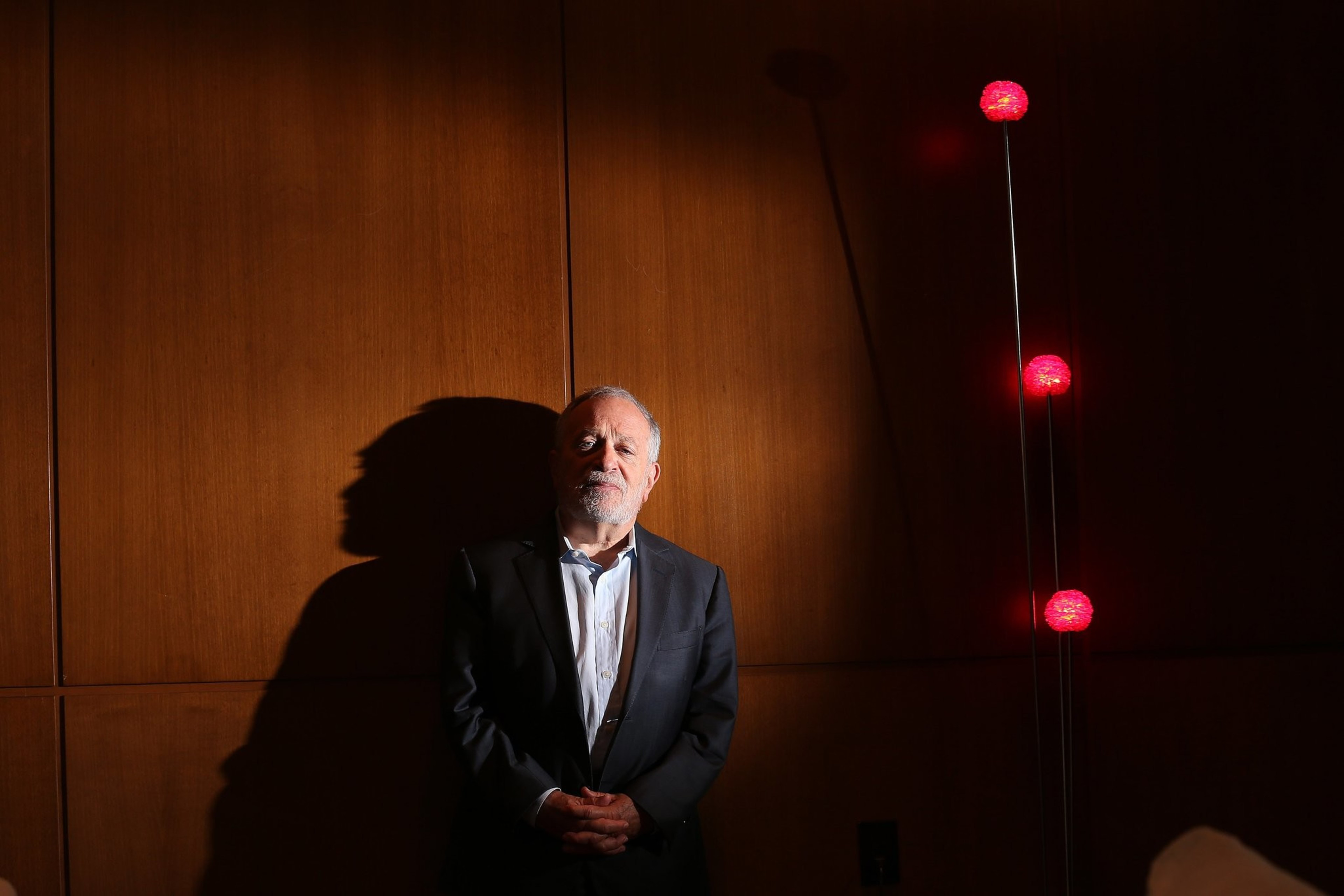 An older man in a dark suit and light shirt stands with hands clasped, casting a shadow on a wooden paneled wall beside three red glowing orbs on tall stands.