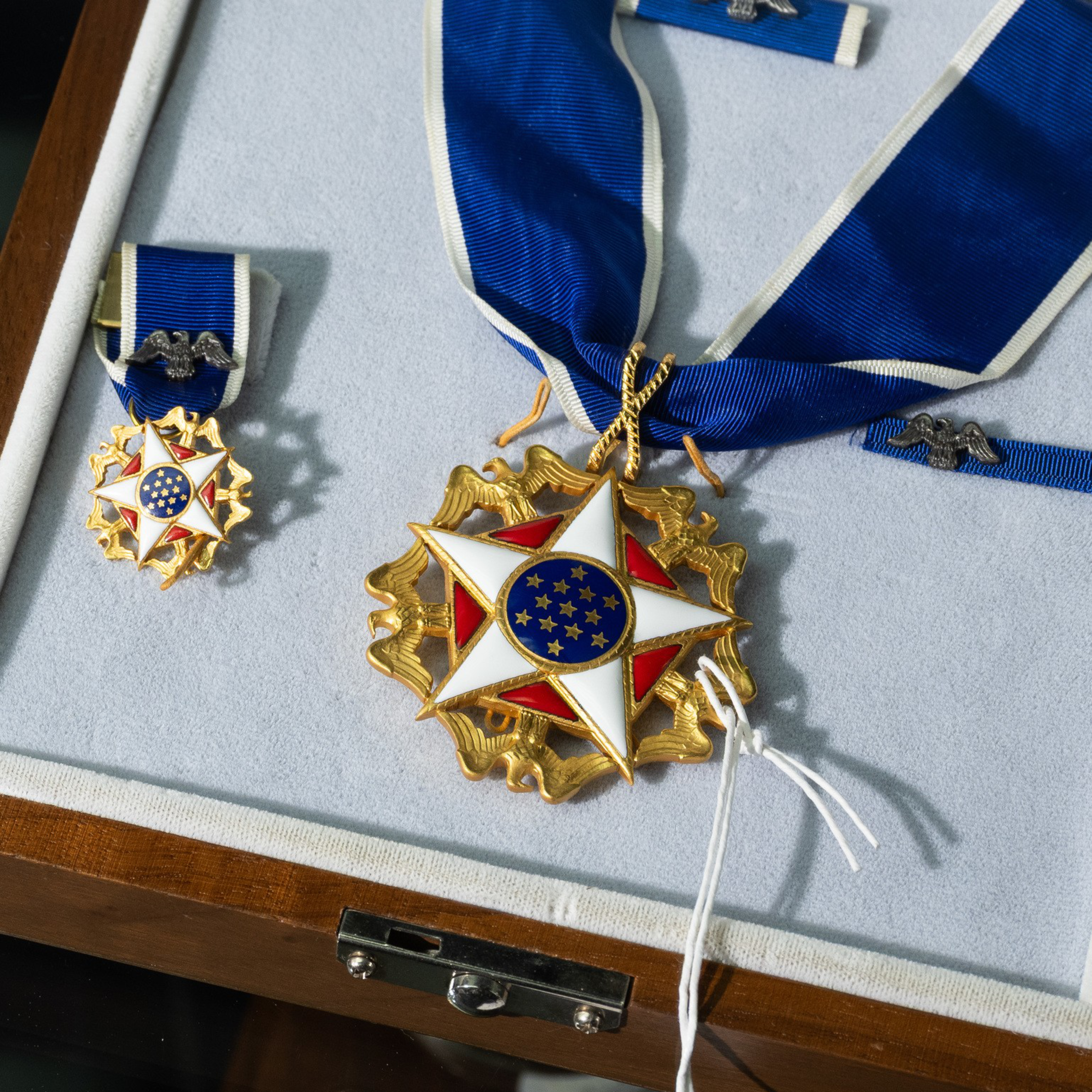 A gold medal with a white and red star, blue circle with stars at its center, attached to a blue ribbon with a small silver eagle clasp, displayed in a case.