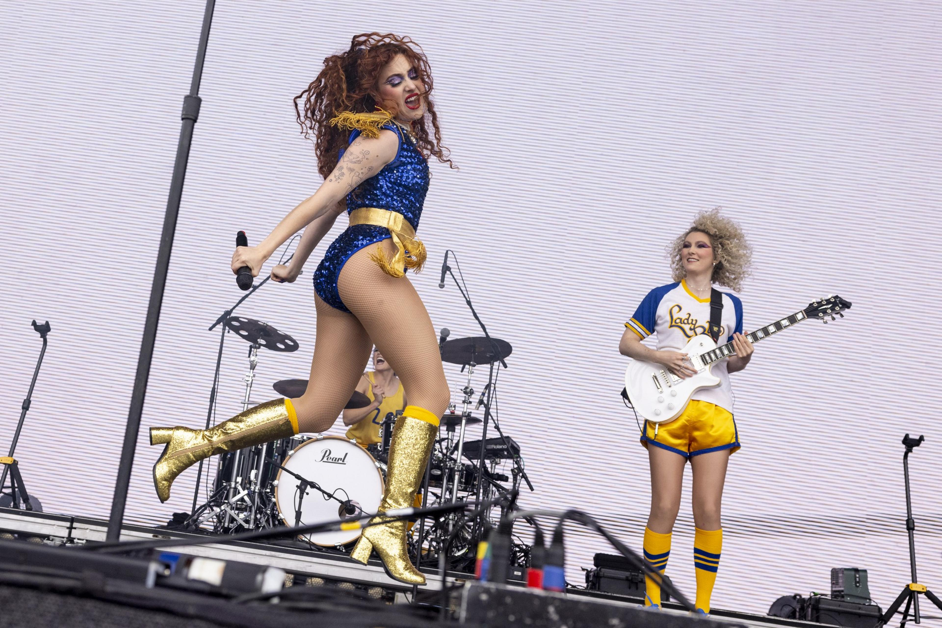 A performer in a sequined blue and gold outfit is energetically dancing and singing on stage, while another musician in a sports jersey and shorts plays a white electric guitar.