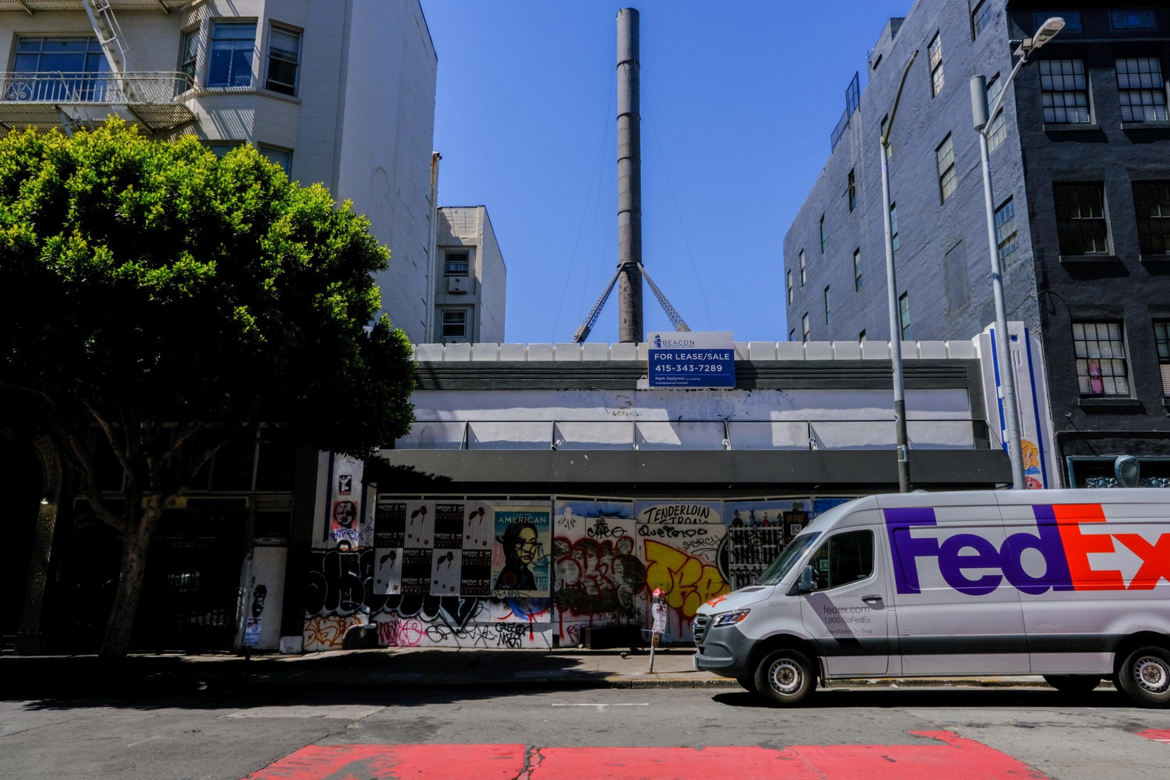 A graffiti-covered storefront with a "For Lease/Sale" sign above it, flanked by buildings and a FedEx delivery van parked on the street.