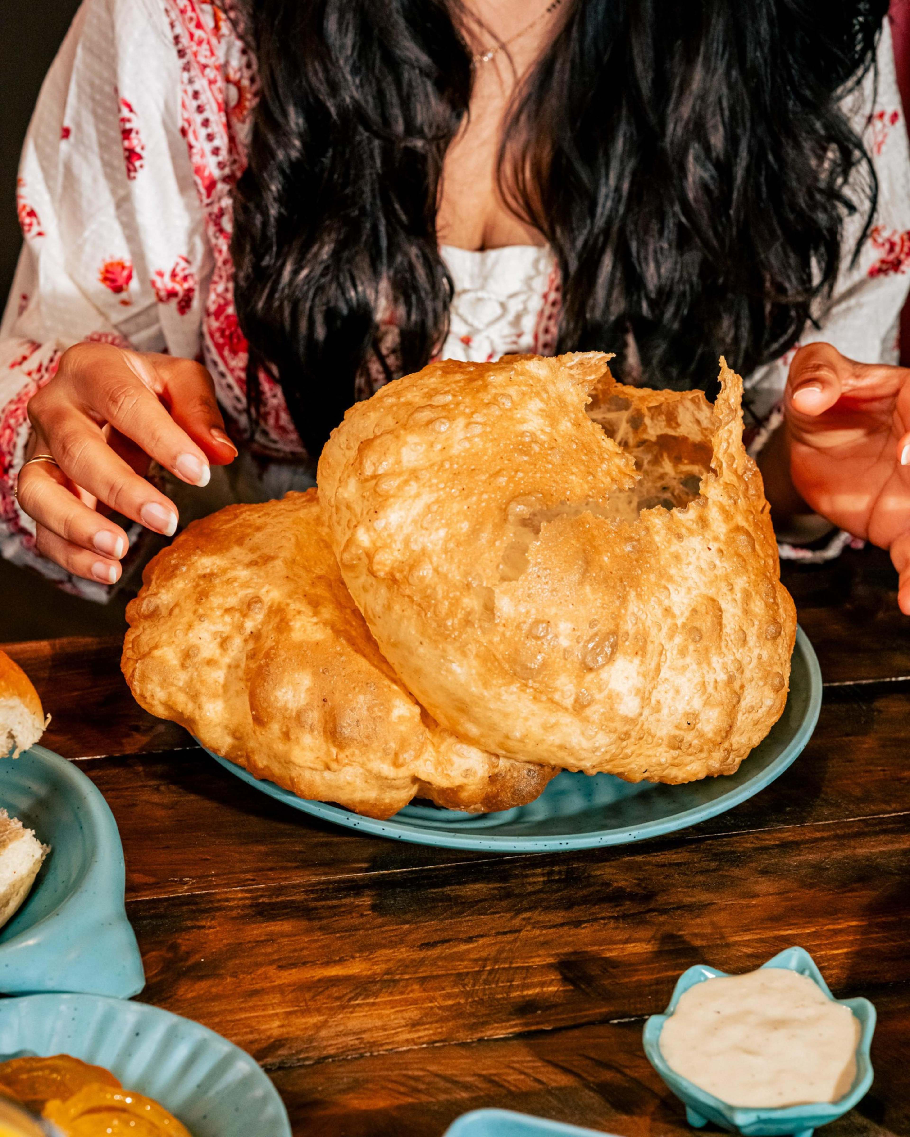 Three large, puffed, golden-brown puris sit on a blue plate, with a person reaching for them, surrounded by small bowls of sauces on a wooden table.