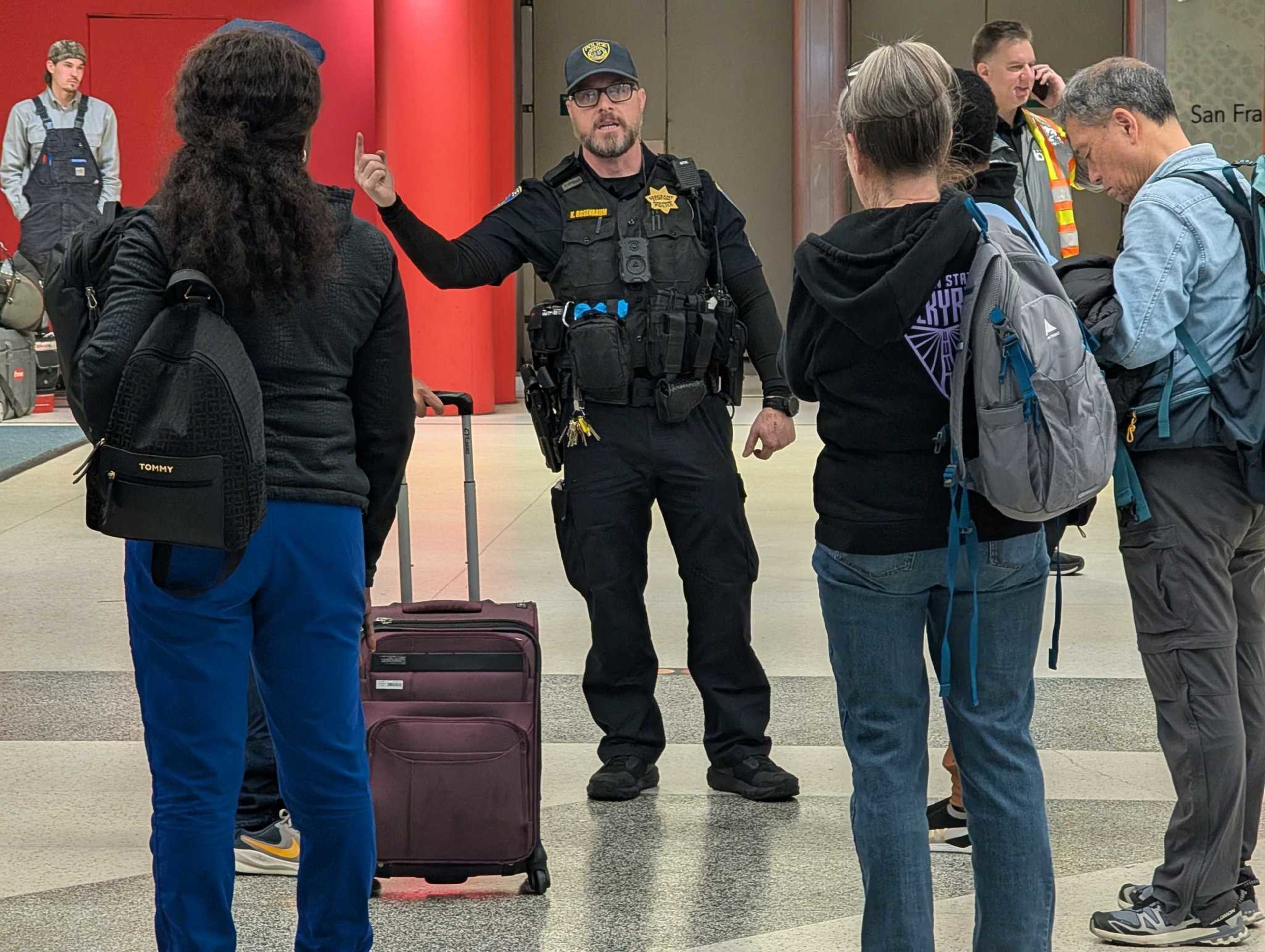 A police officer is speaking to a small group. One person has a suitcase and a backpack. Others are holding backpacks, and one uses a phone, with a red wall behind.