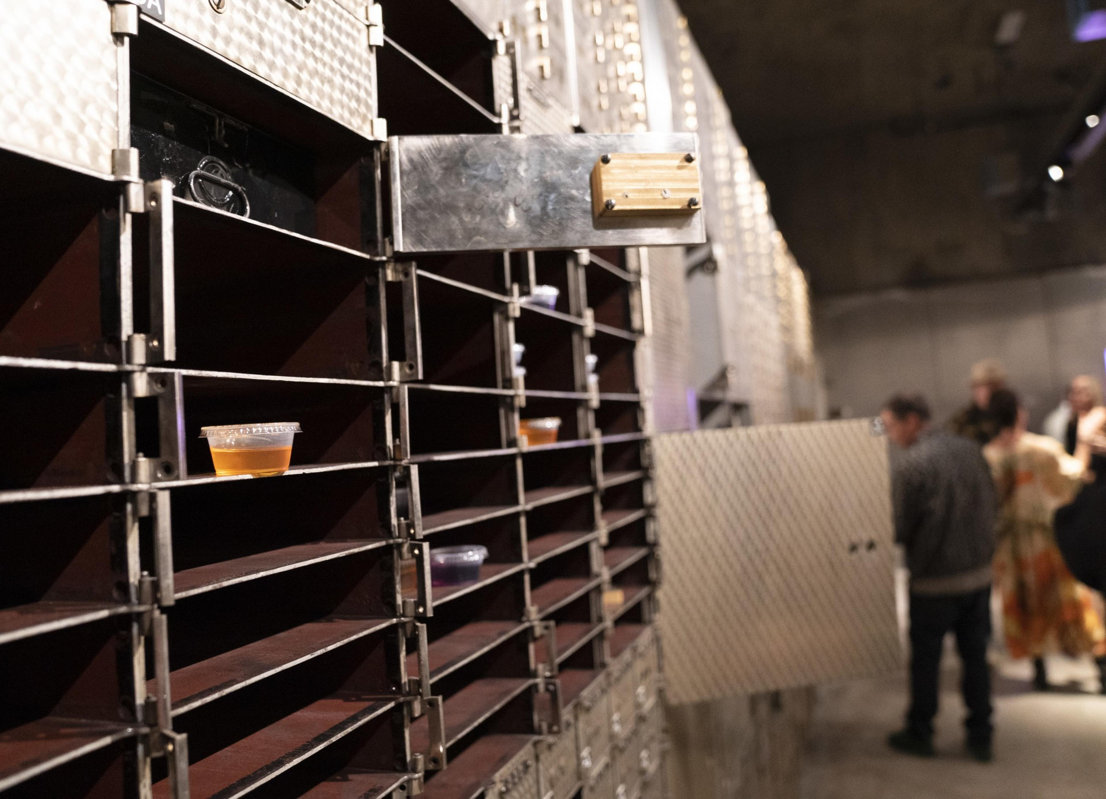 The image shows industrial-style shelves with small cups of liquid on them. In the background, a group of people stand, slightly out of focus.