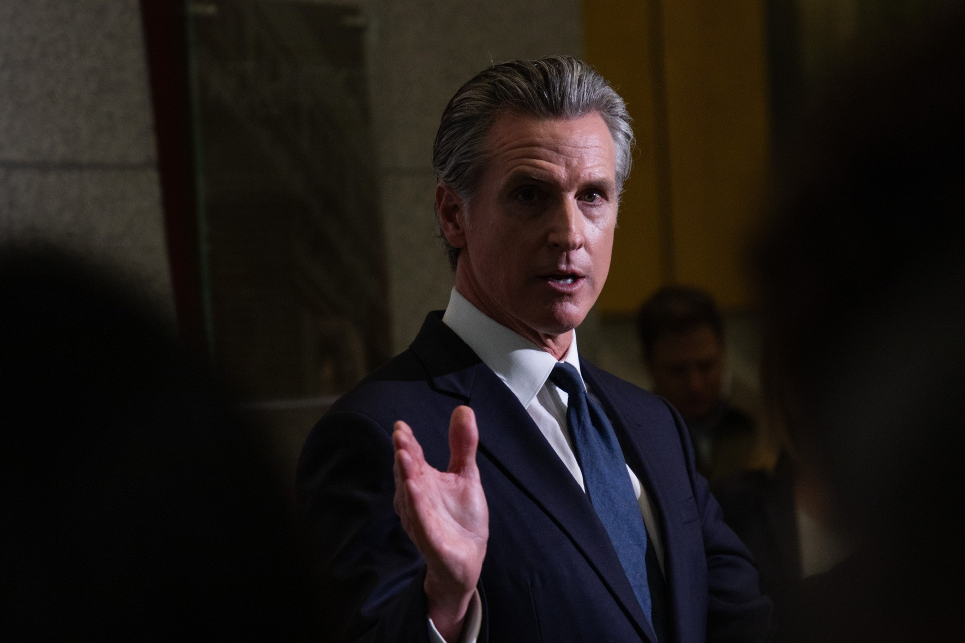 A man in a dark suit gestures with one hand raised slightly. He is standing indoors, lit by soft, focused lighting with a few blurred people in the background.
