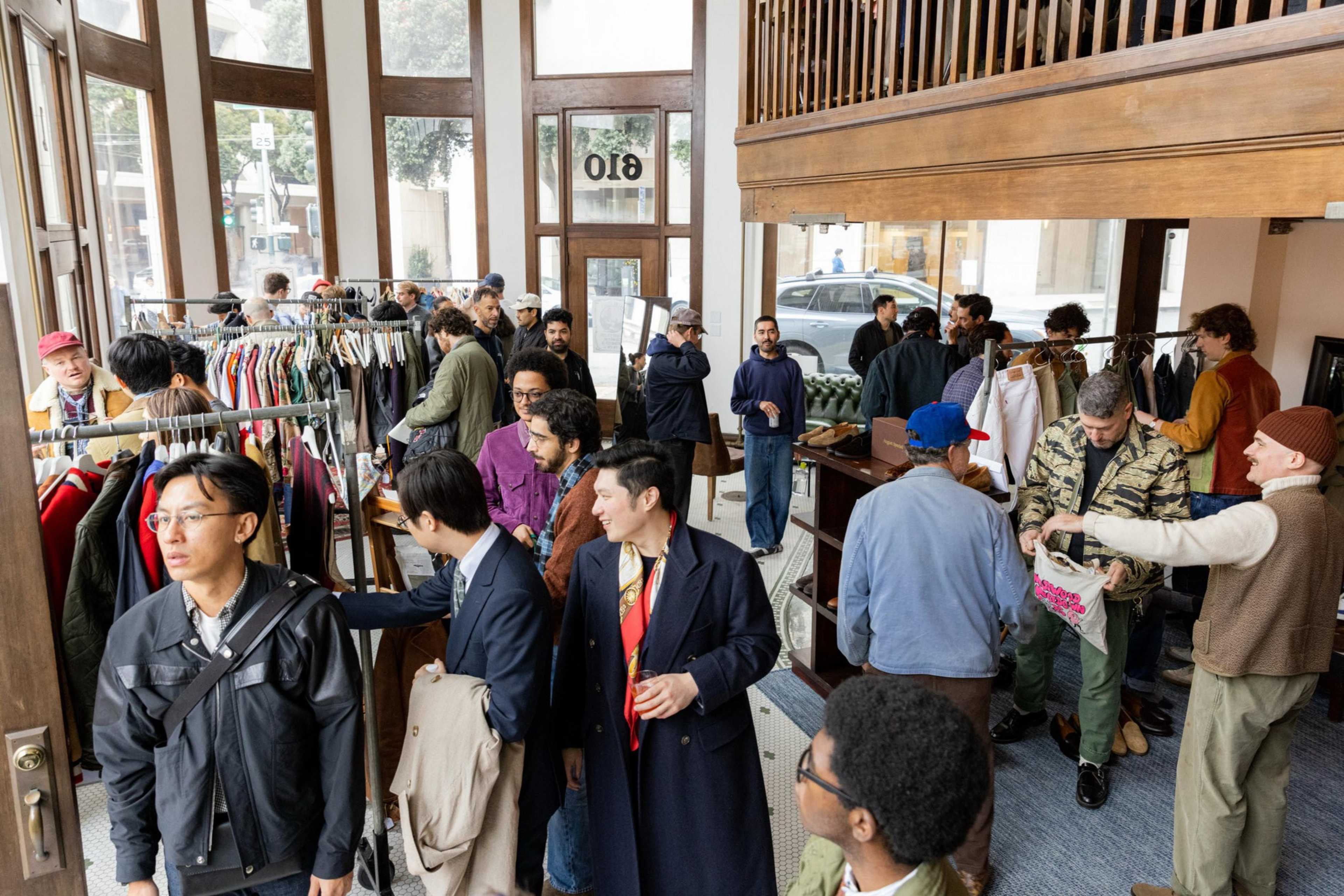 A crowded clothing store with people browsing racks, chatting, and trying on clothes near large windows and a wooden balcony above.