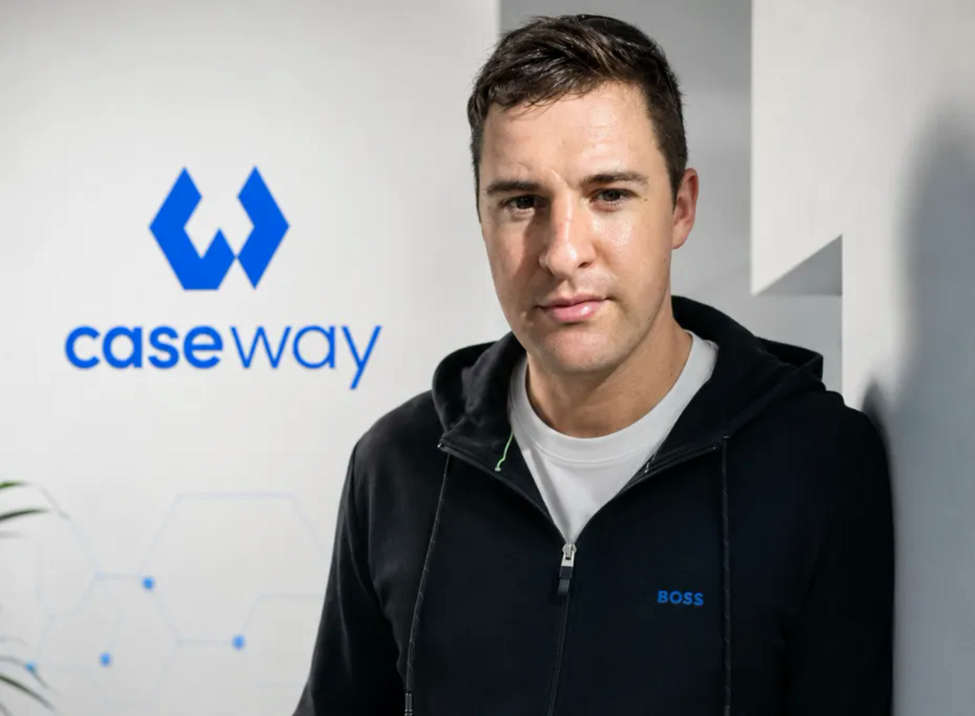 A man in a black Boss hoodie and white shirt stands beside a wall with a blue "case way" logo and geometric designs.