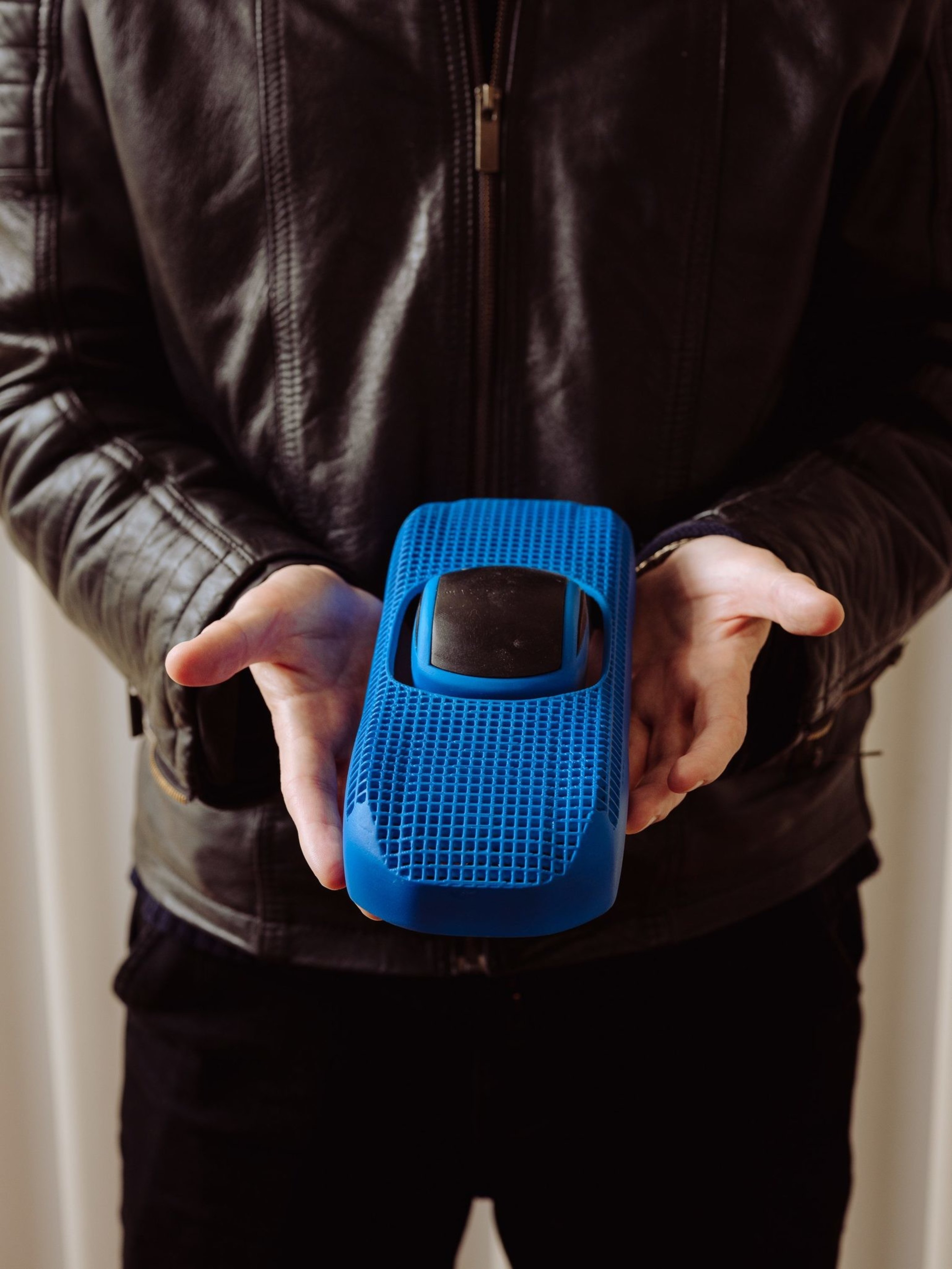 A person in a leather jacket holds a small blue object resembling a futuristic remote or gadget with a grid texture, centered in their open palms.