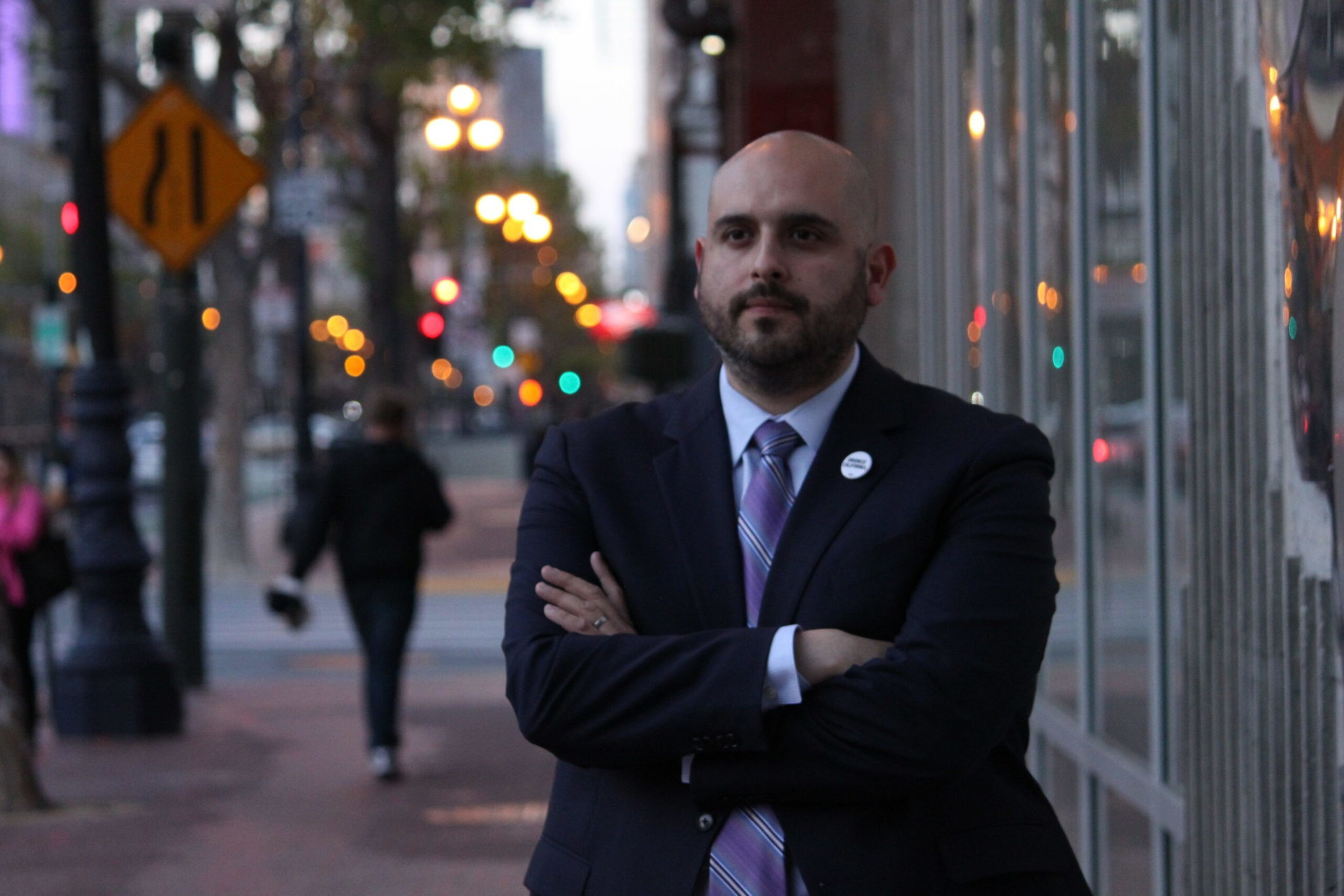 A bald man with a beard in a dark suit and striped tie stands with arms crossed on a city sidewalk at dusk, blurred streetlights glowing behind him.