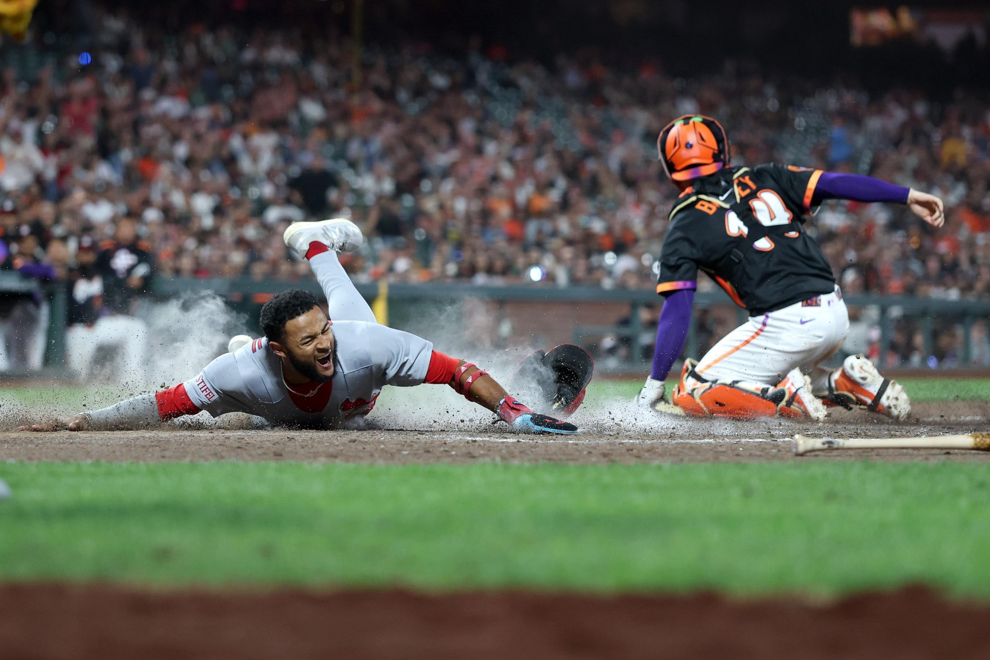 A baseball player in a gray uniform slides headfirst into home plate while a catcher in a black and orange uniform attempts to tag him out.