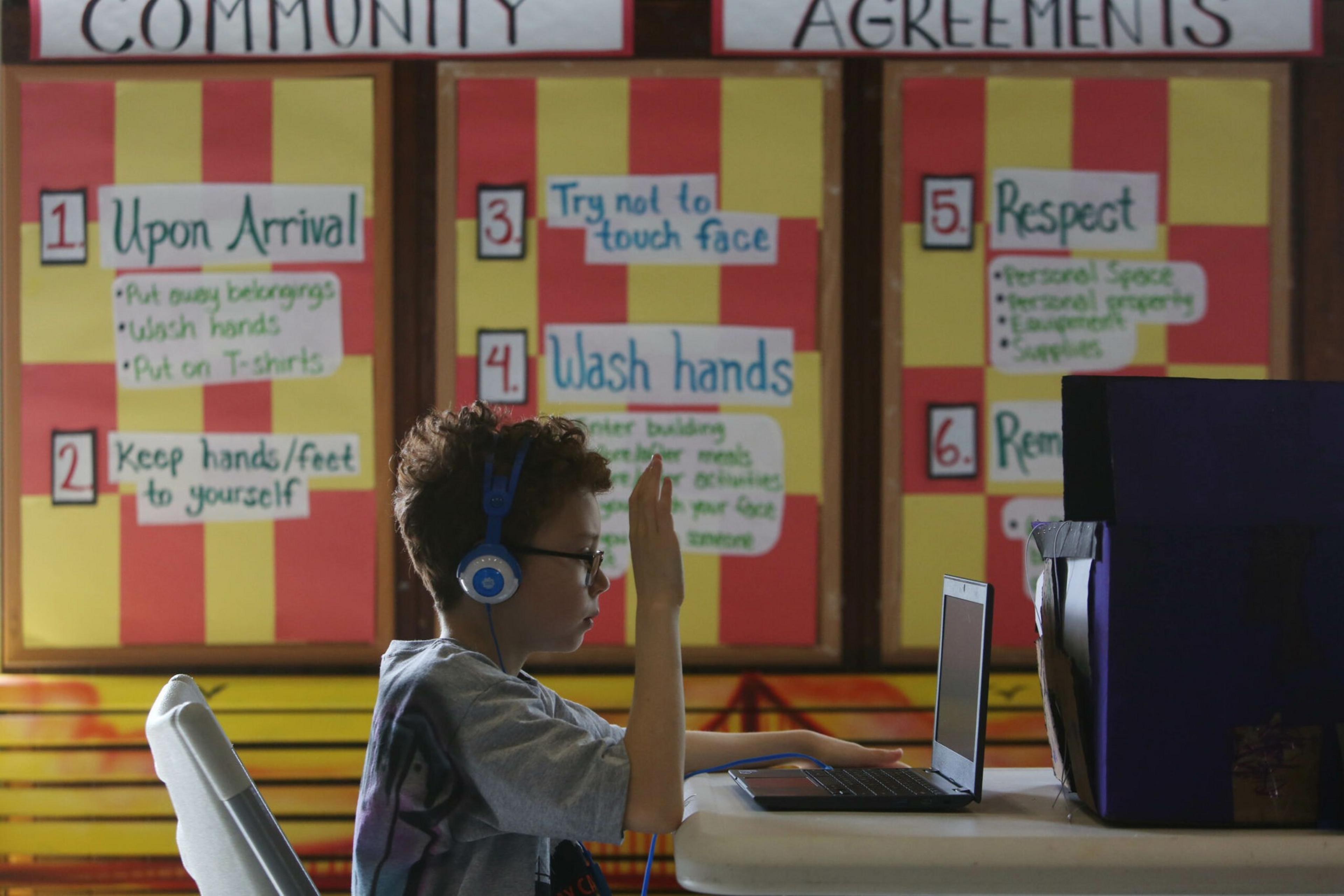 A child wearing blue headphones sits at a table, raising one hand while looking at a laptop, with colorful community agreement posters on the wall behind.