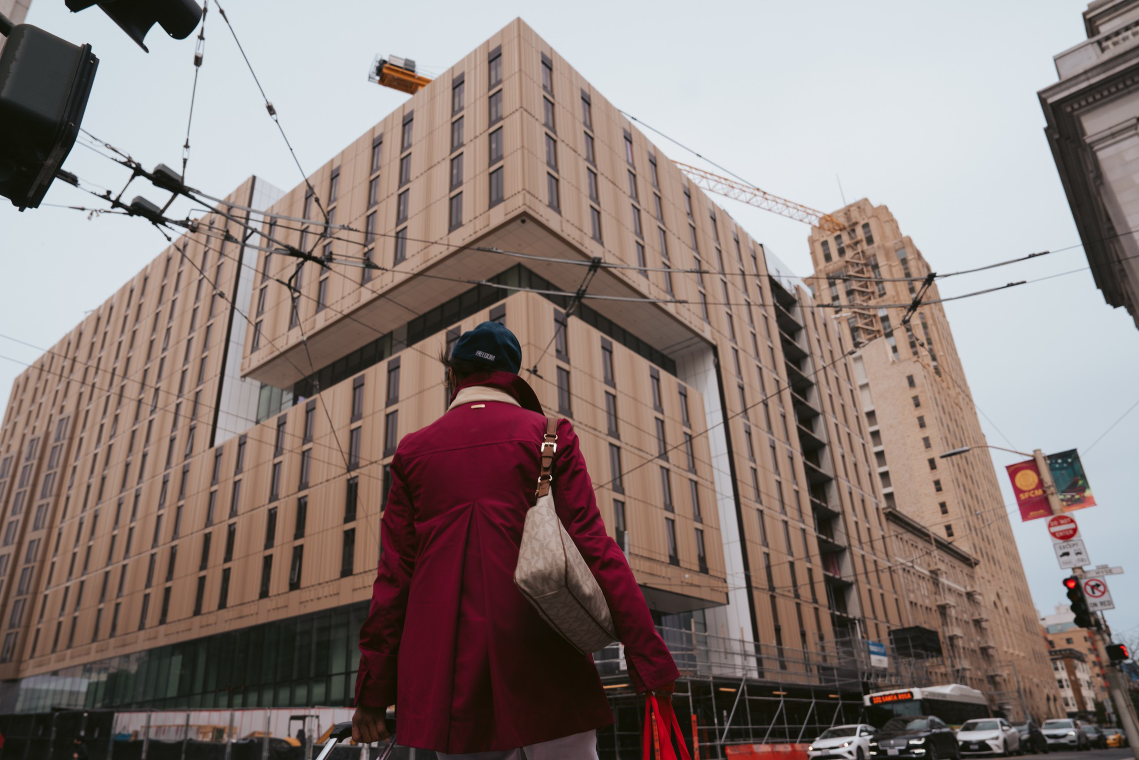A person in a red coat faces modern buildings; wires and street signs crisscross above.