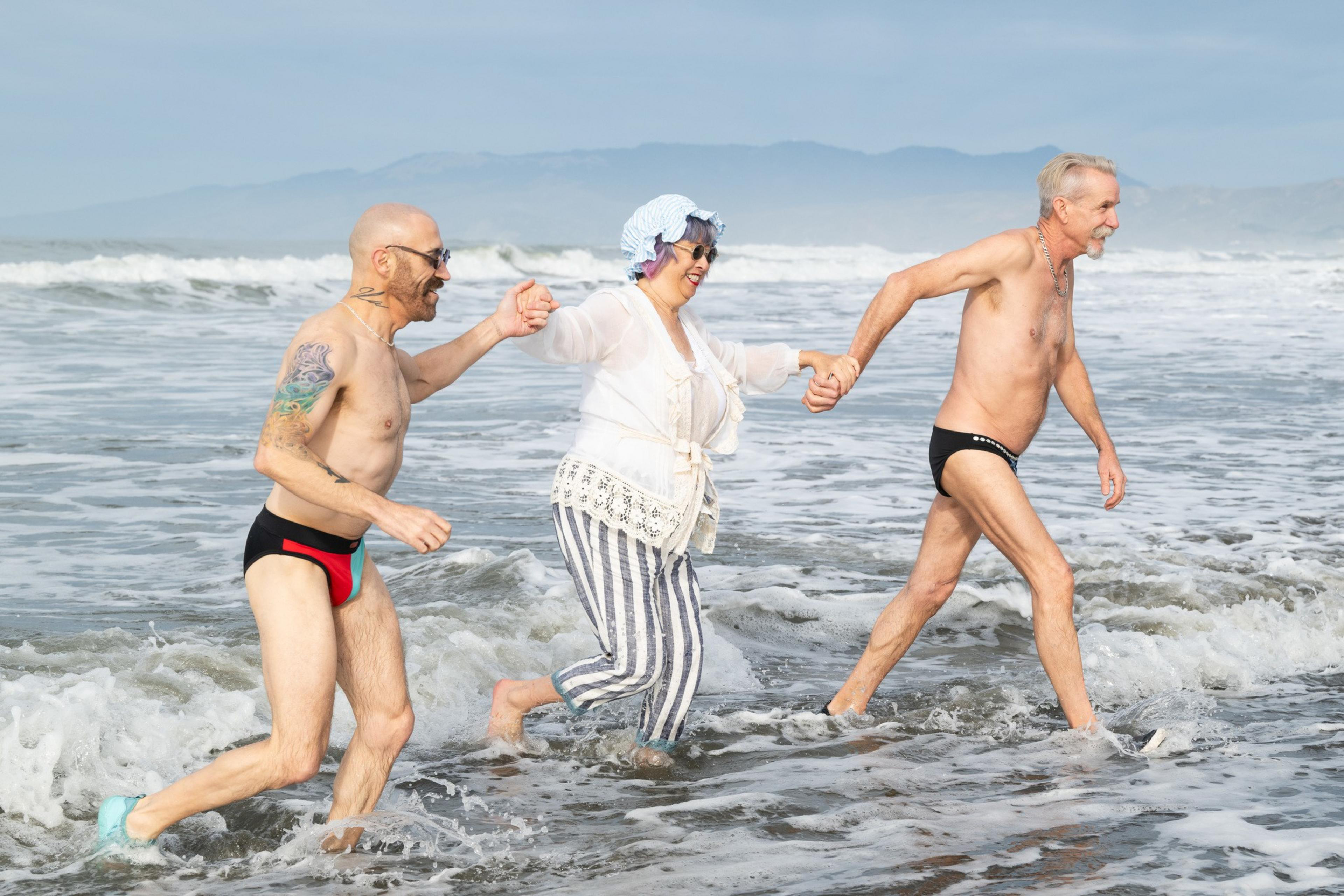 Three people joyfully run hand-in-hand through shallow ocean waves, two wearing swimsuits and one in a hat with striped pants, against a distant mountain backdrop.