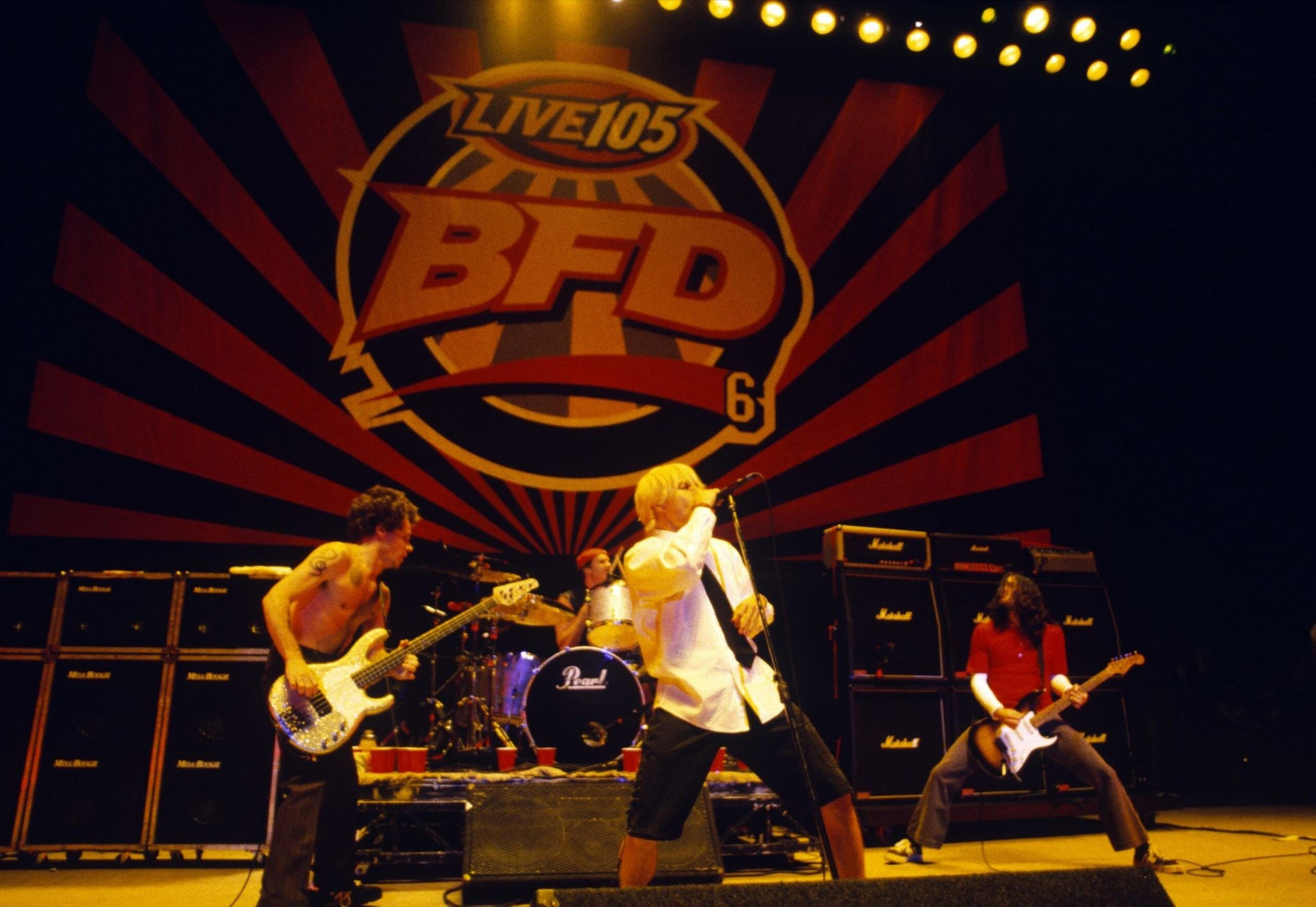 A rock band performs on stage with guitarists, a drummer, and a vocalist under a "LIVE 105 BFD" banner and bright yellow stage lights.