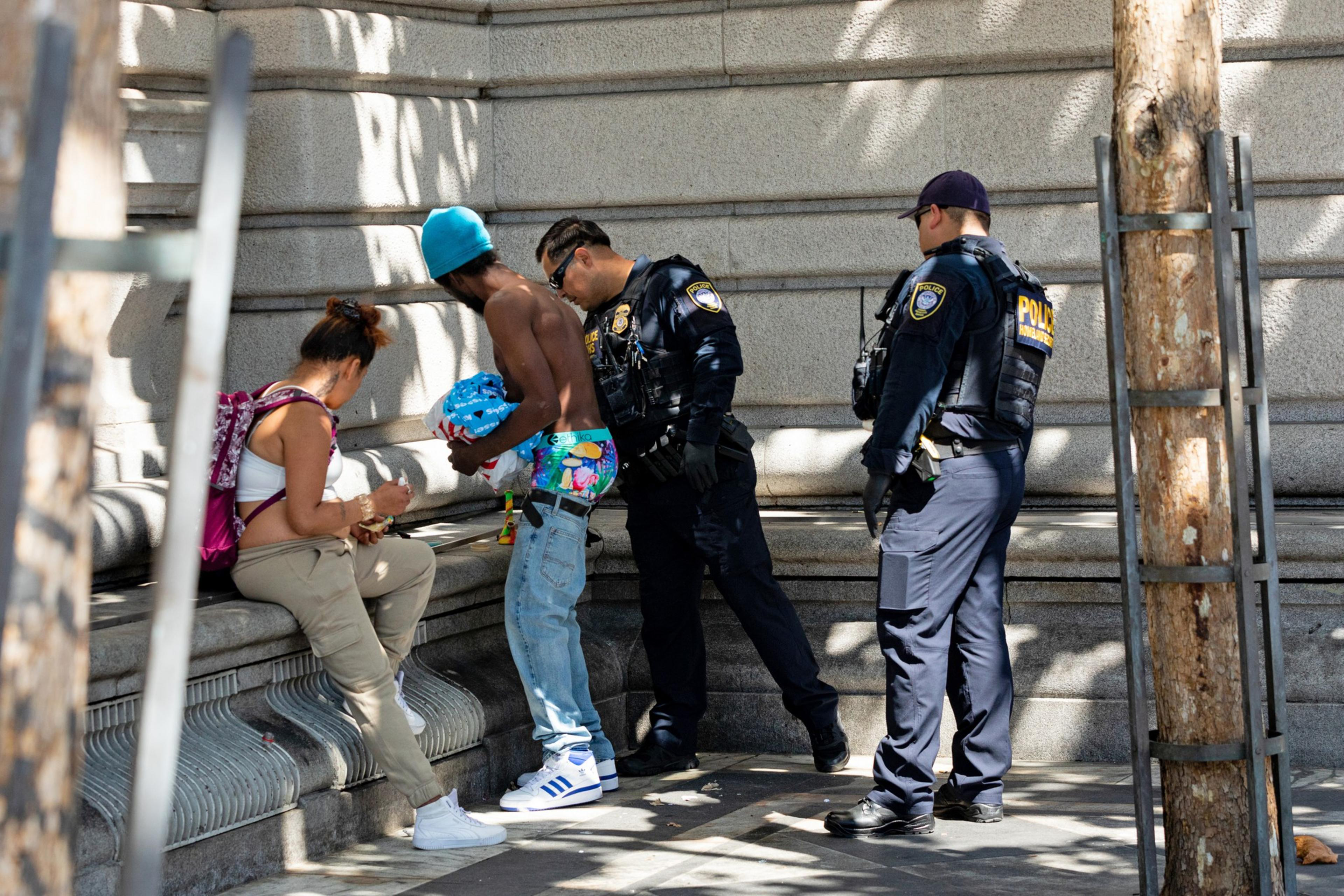 Two police officers are interacting with a seated woman and a standing shirtless man wearing a blue beanie and colorful pants in an urban setting.