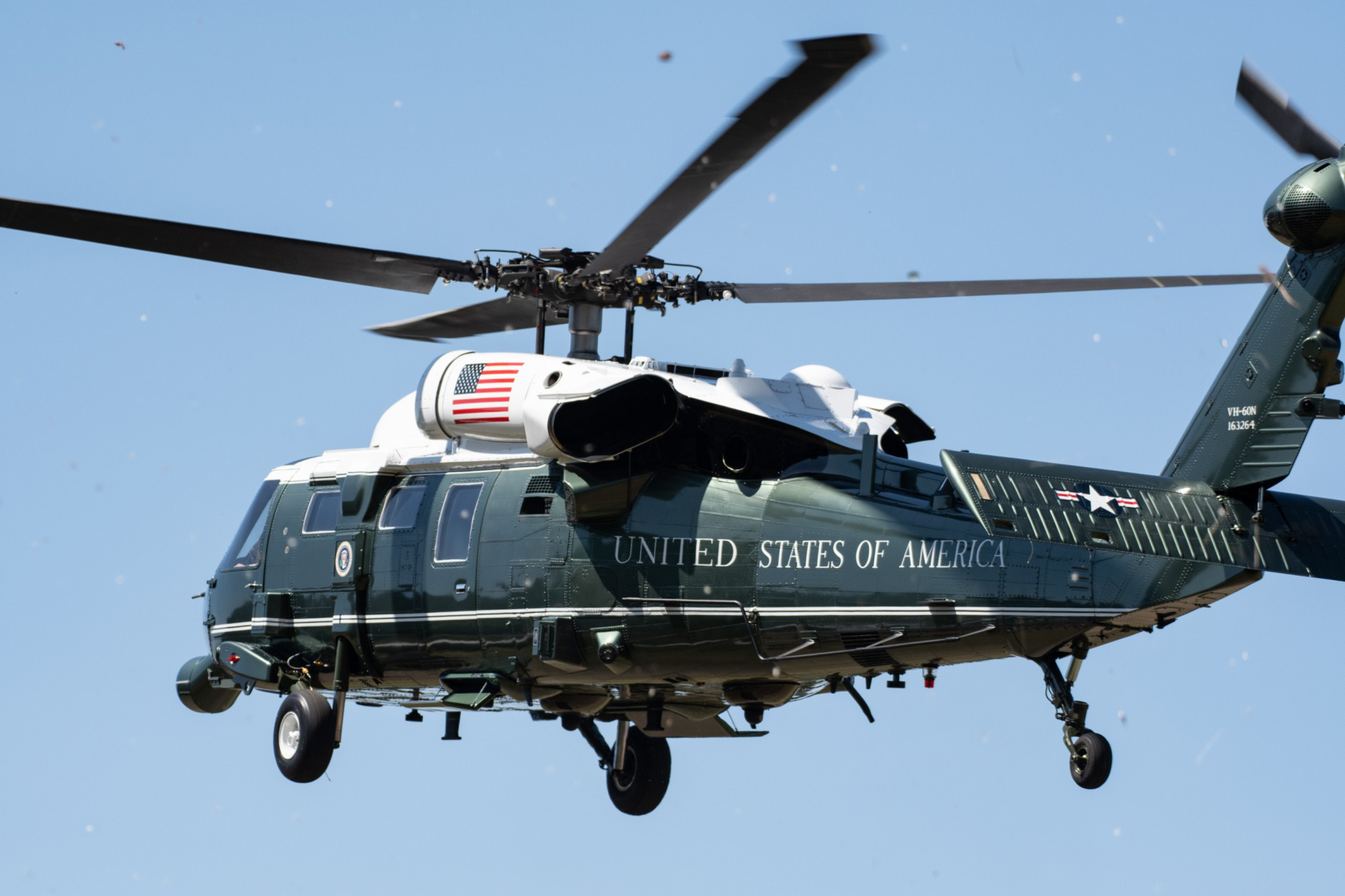 A dark green United States helicopter with the American flag and "UNITED STATES OF AMERICA" on its side flies against a clear blue sky.