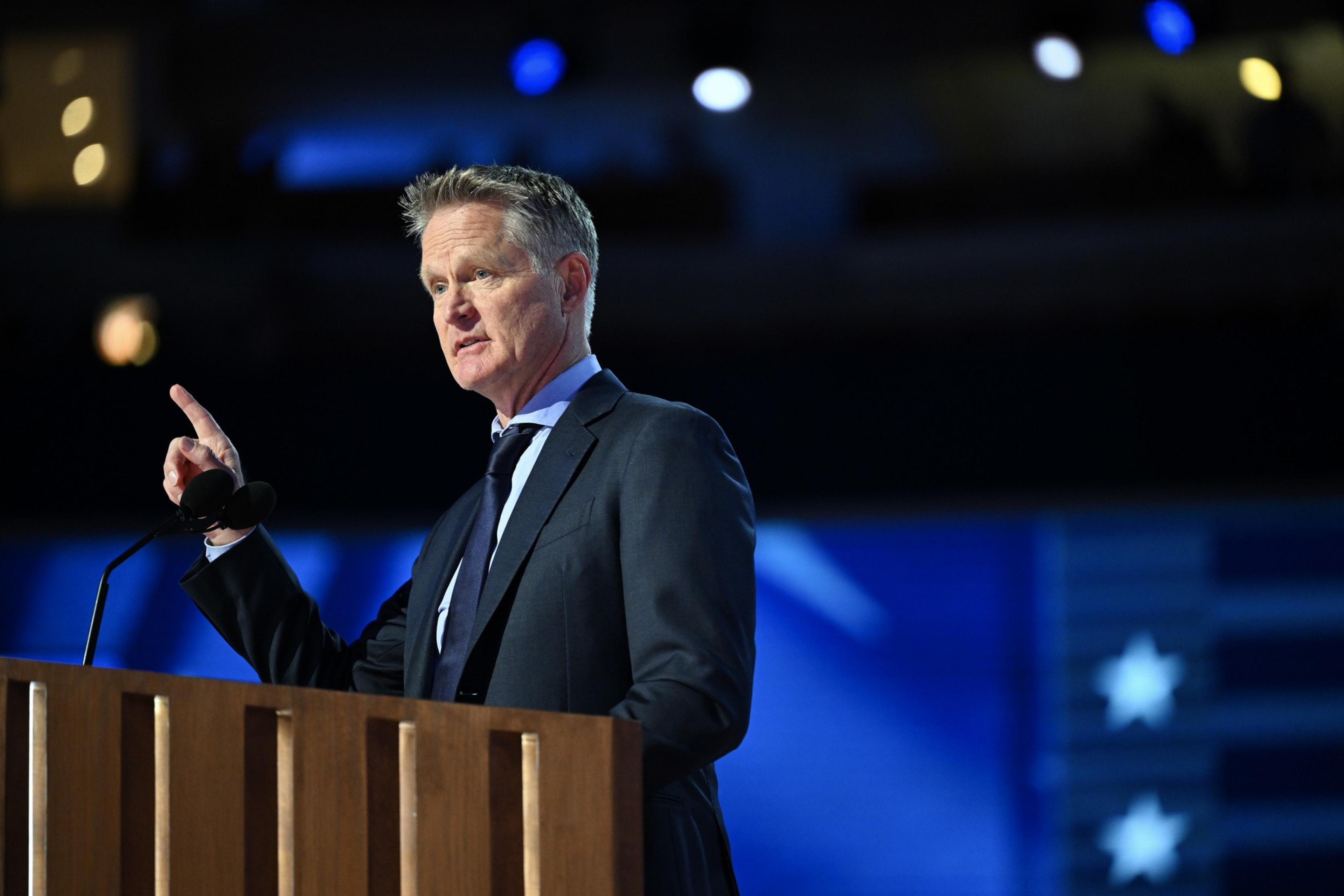 A man in a suit gestures while speaking at a wooden podium with a microphone, set against a blue-toned, blurred background featuring stars.