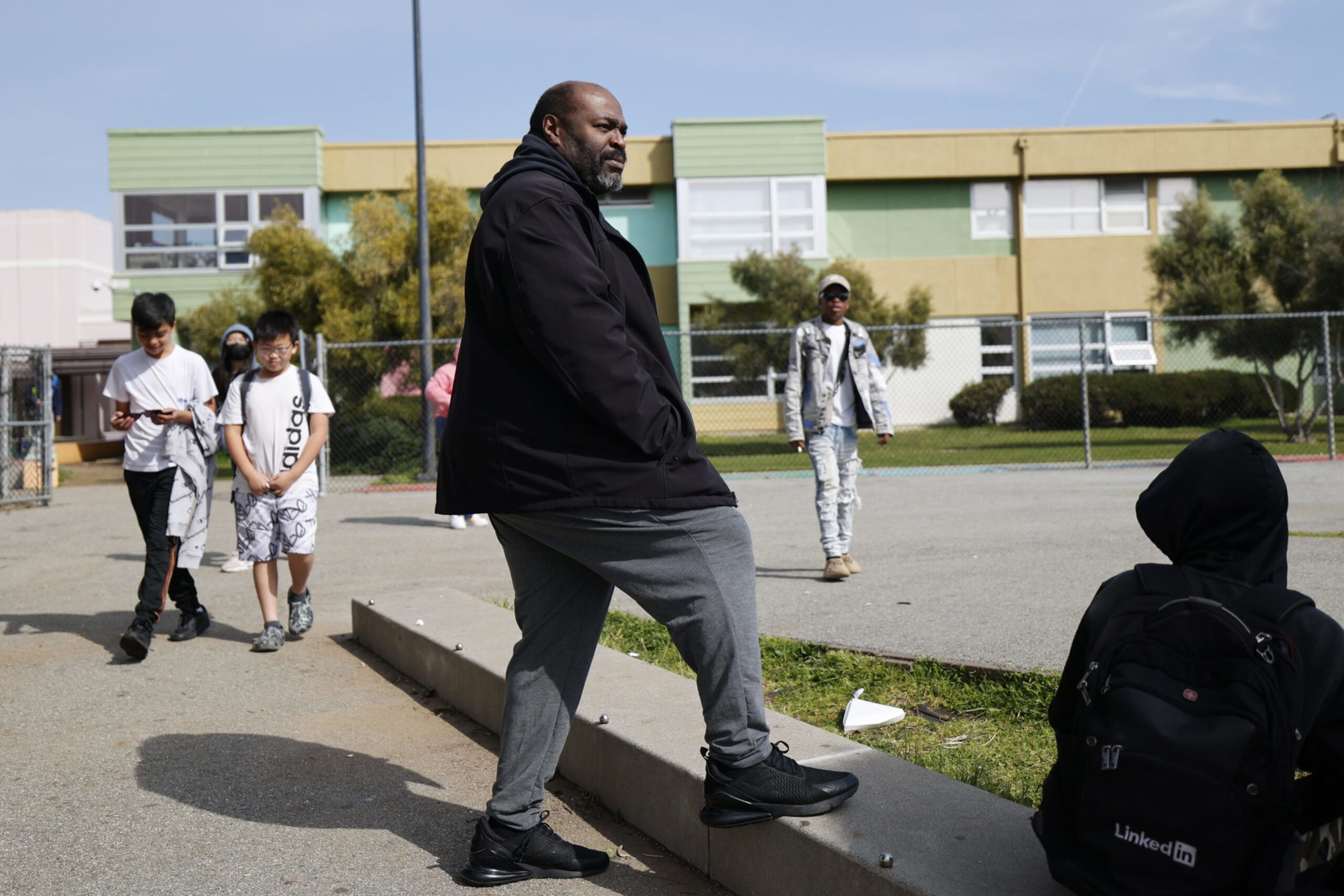 A man in a black jacket and gray pants leans on a curb while several children walk nearby in a schoolyard with colorful buildings in the background.