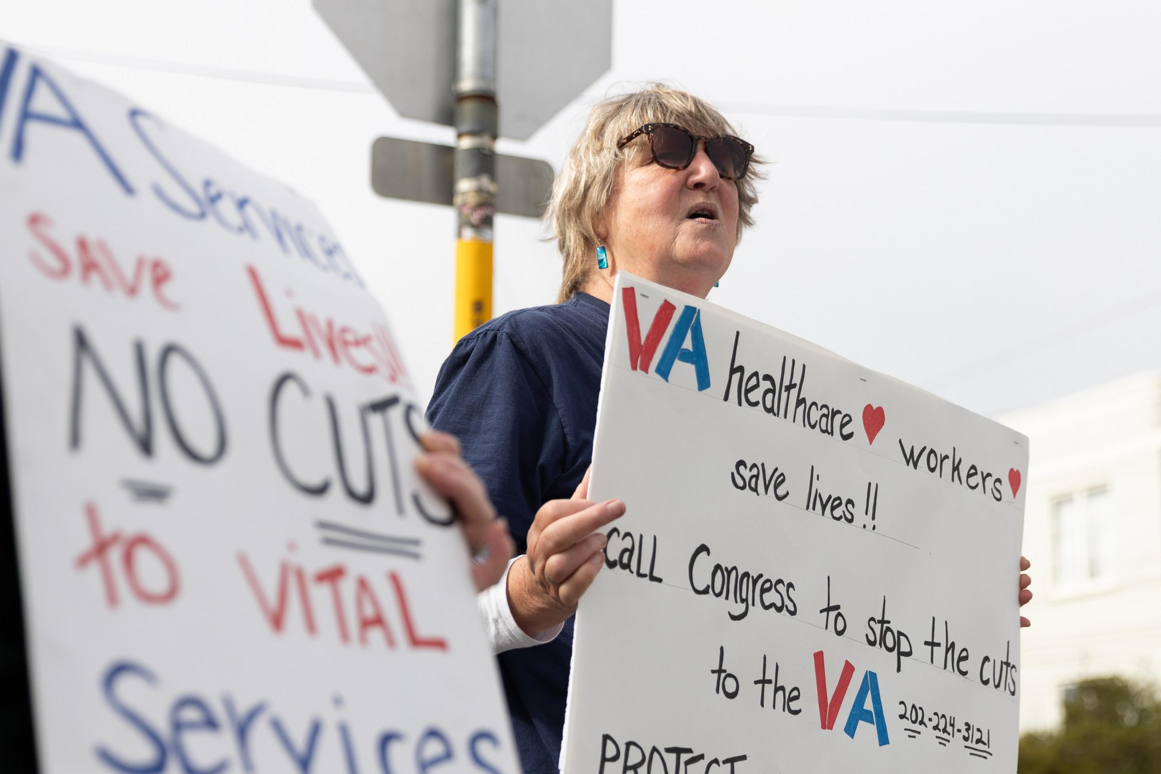 A person holds signs protesting cuts to VA healthcare services, emphasizing their importance in saving lives.