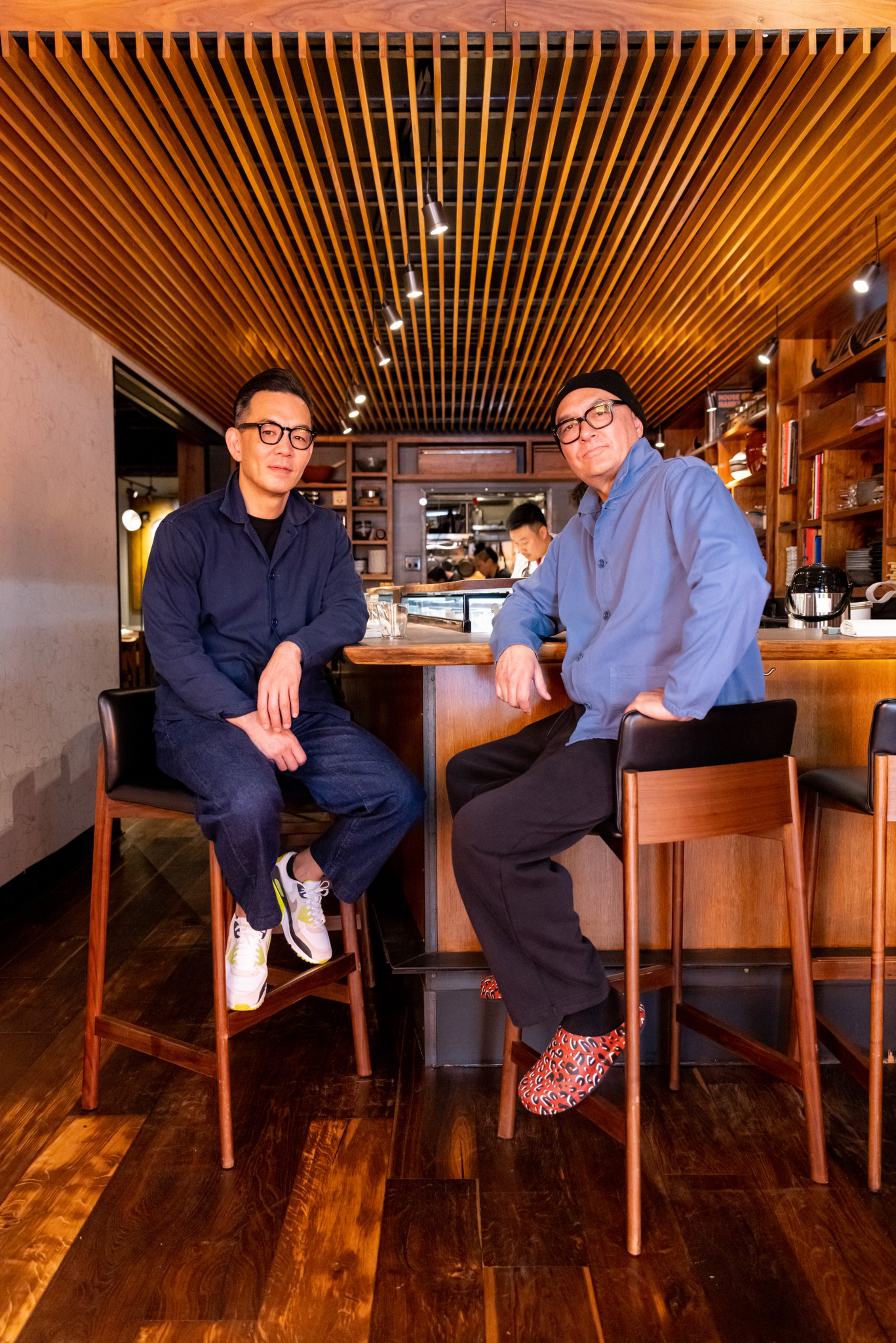 Two men in glasses sit on tall wooden stools at a bar with a wooden slatted ceiling, warm lighting, and shelves filled with items.