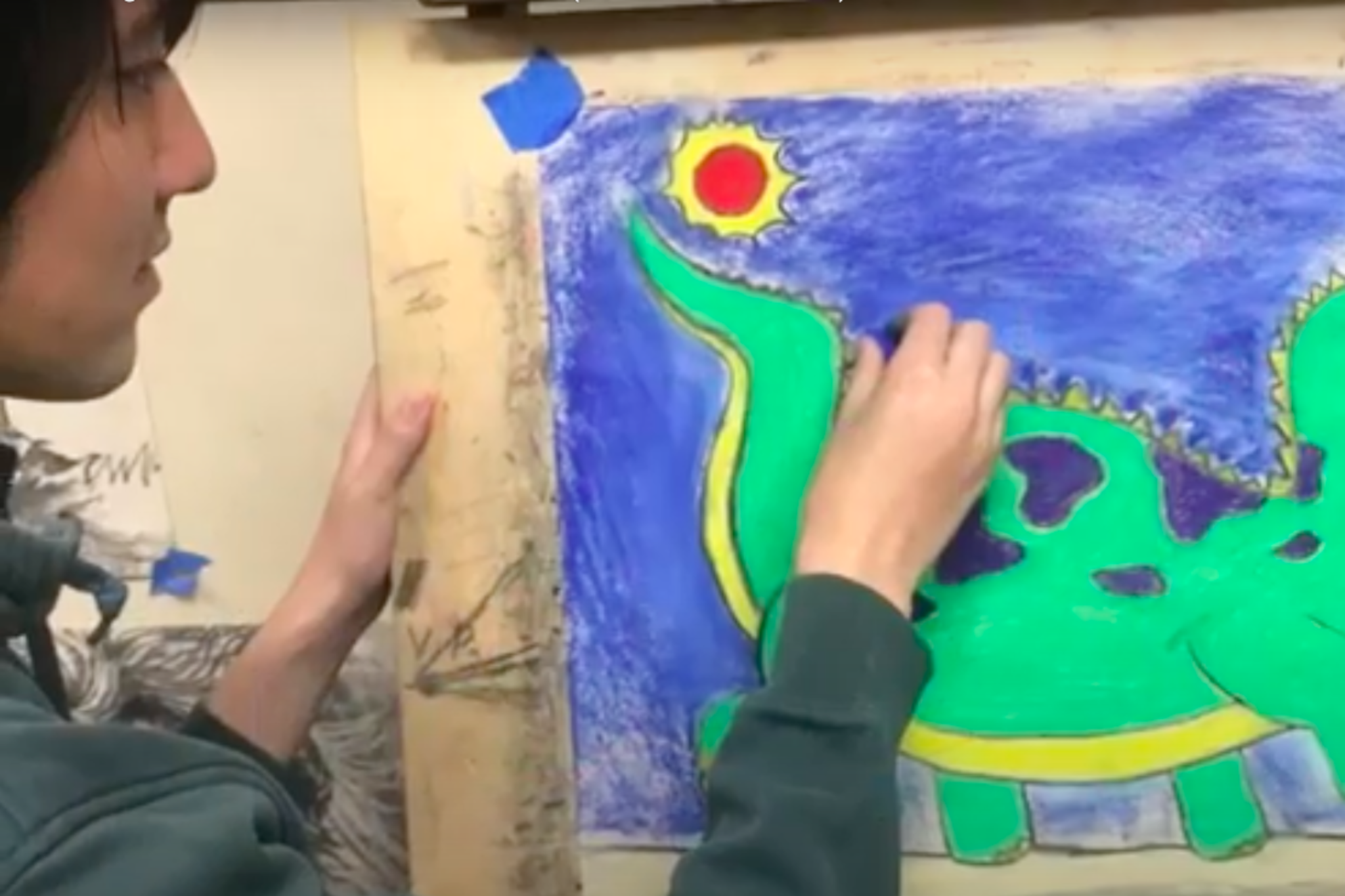 A person colors a green dinosaur with purple spots and yellow accents against a blue background with a red sun in the top left corner.
