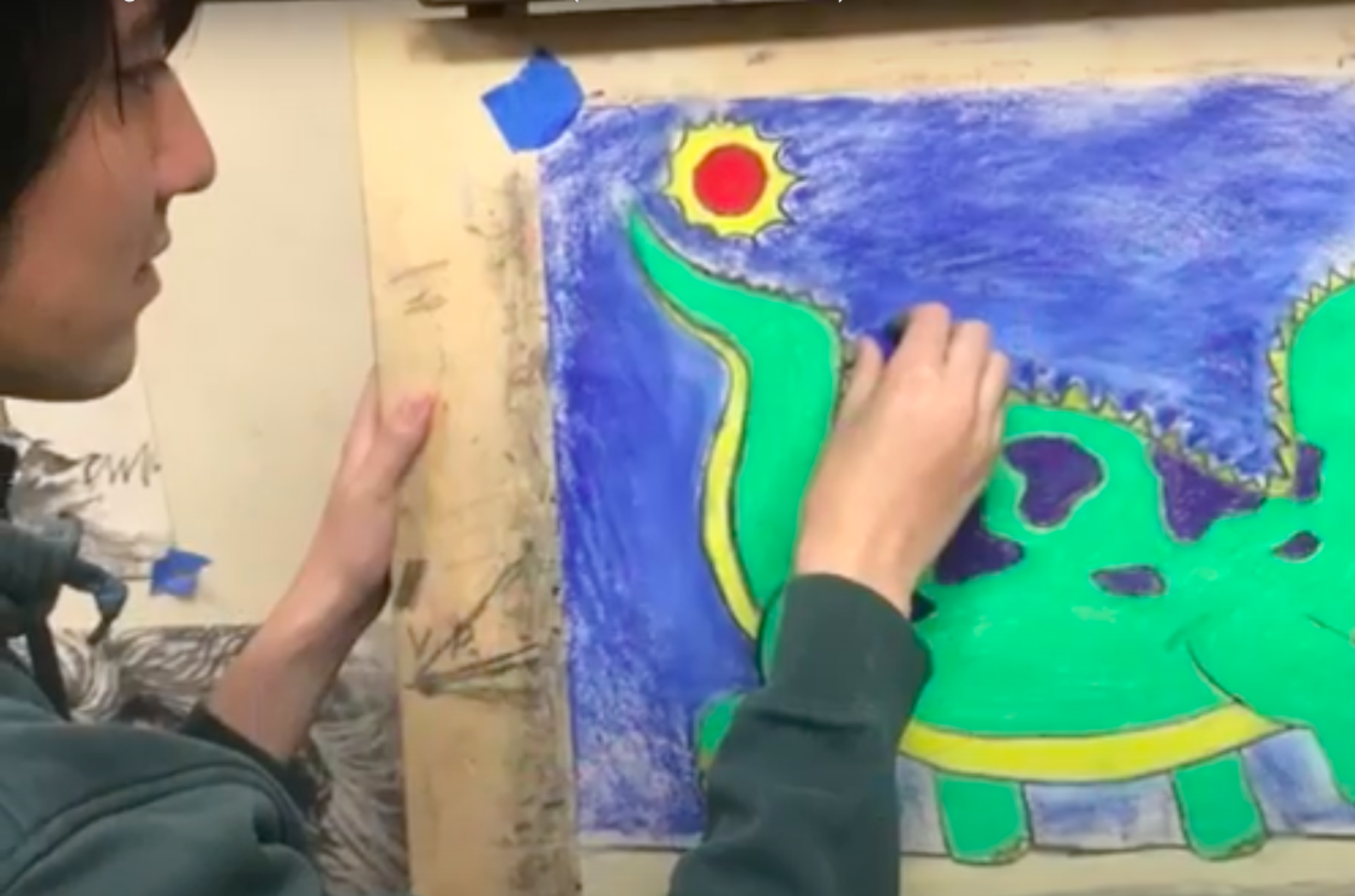 A person colors a green dinosaur with purple spots and yellow accents against a blue background with a red sun in the top left corner.
