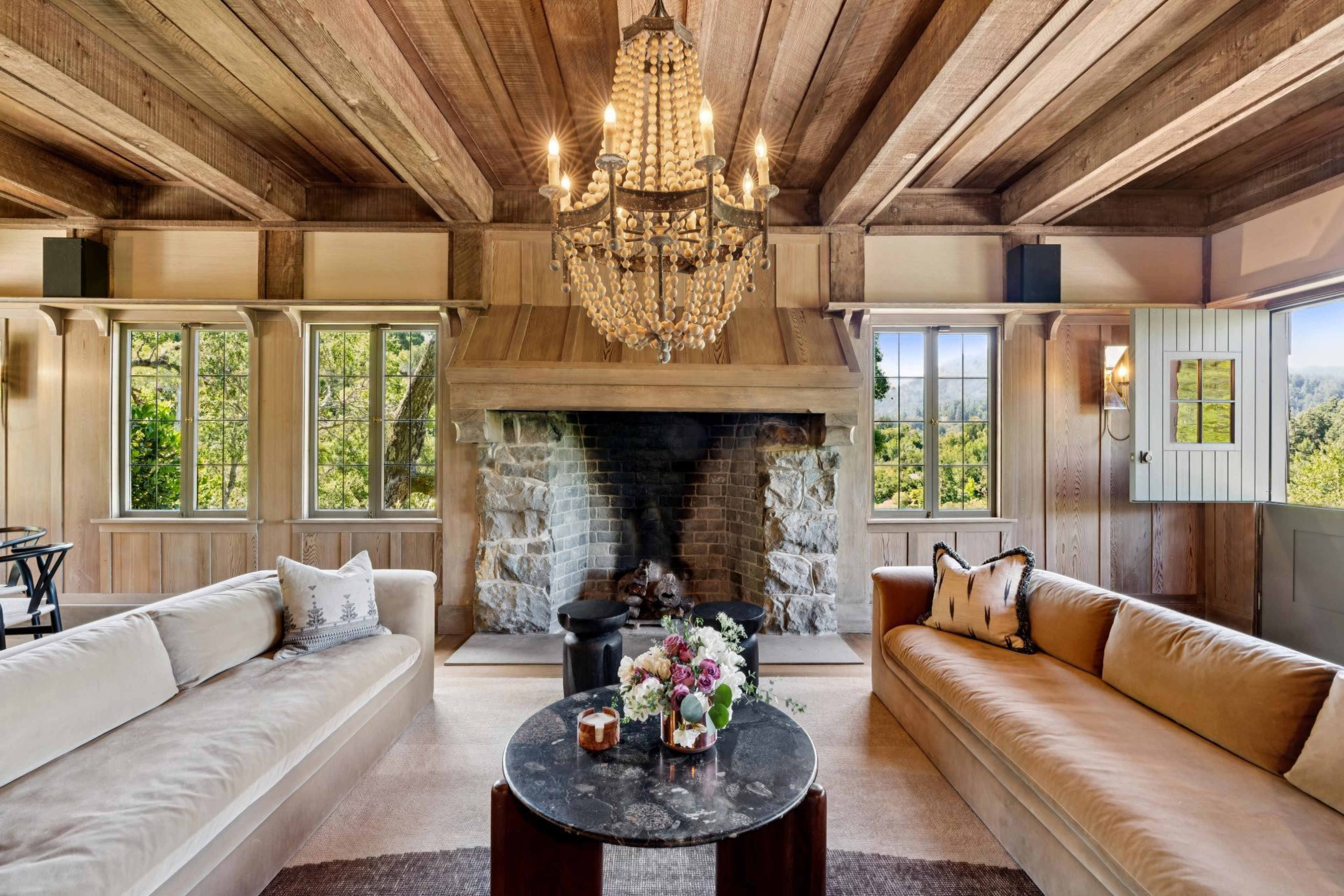 A rustic living room with wooden walls and ceiling, a stone fireplace, two long sofas, a round black coffee table, and a chandelier hanging above.
