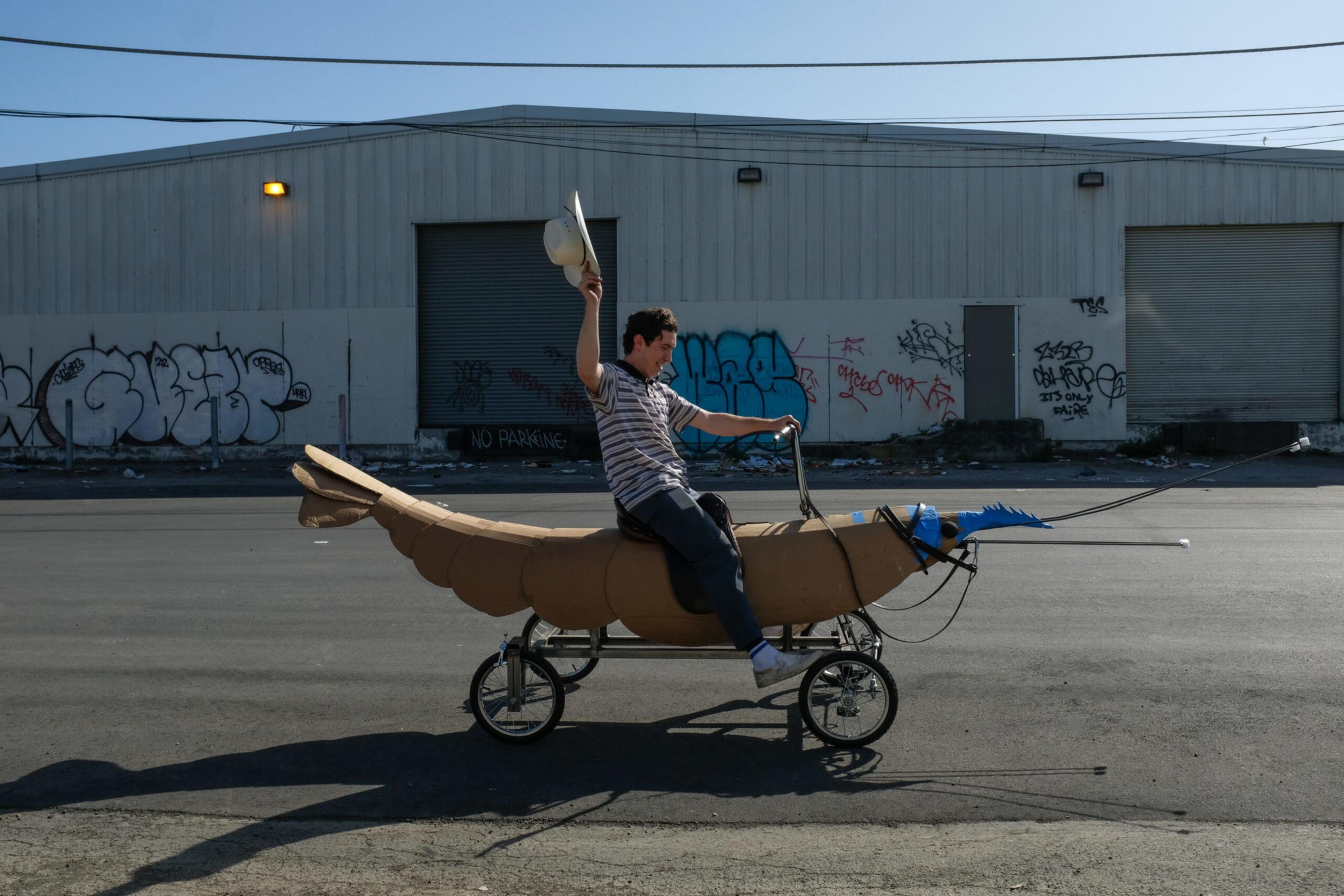 A man rides a shrimp-shaped vehicle on a street, holding a hat in the air, with graffiti-covered industrial buildings in the background.