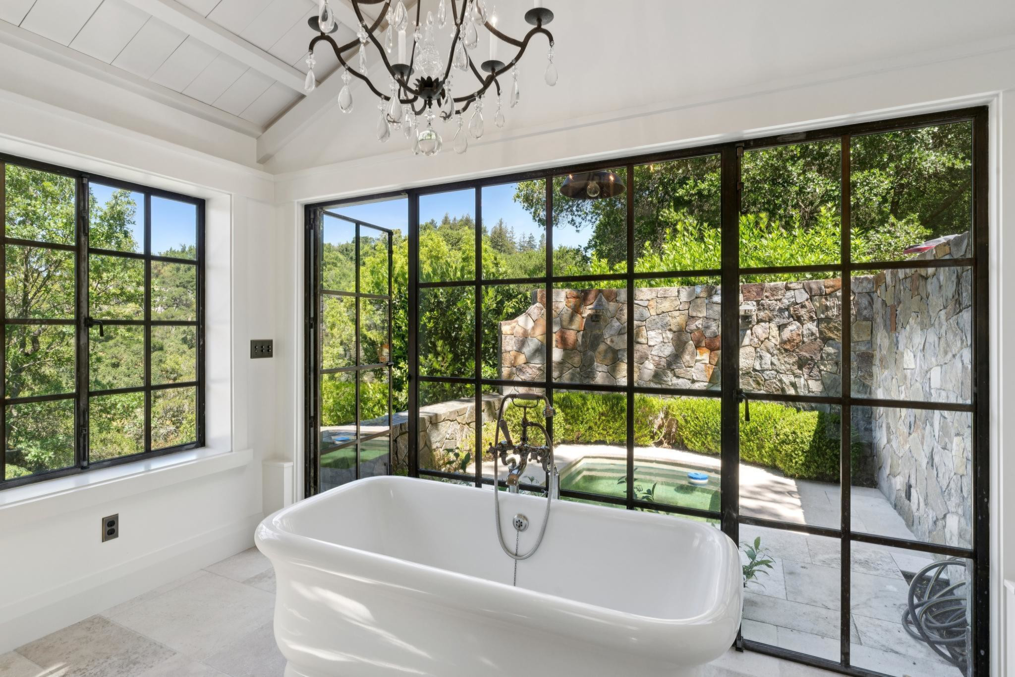 A white clawfoot bathtub faces large black-framed windows overlooking a stone-walled garden with greenery and a small pool.