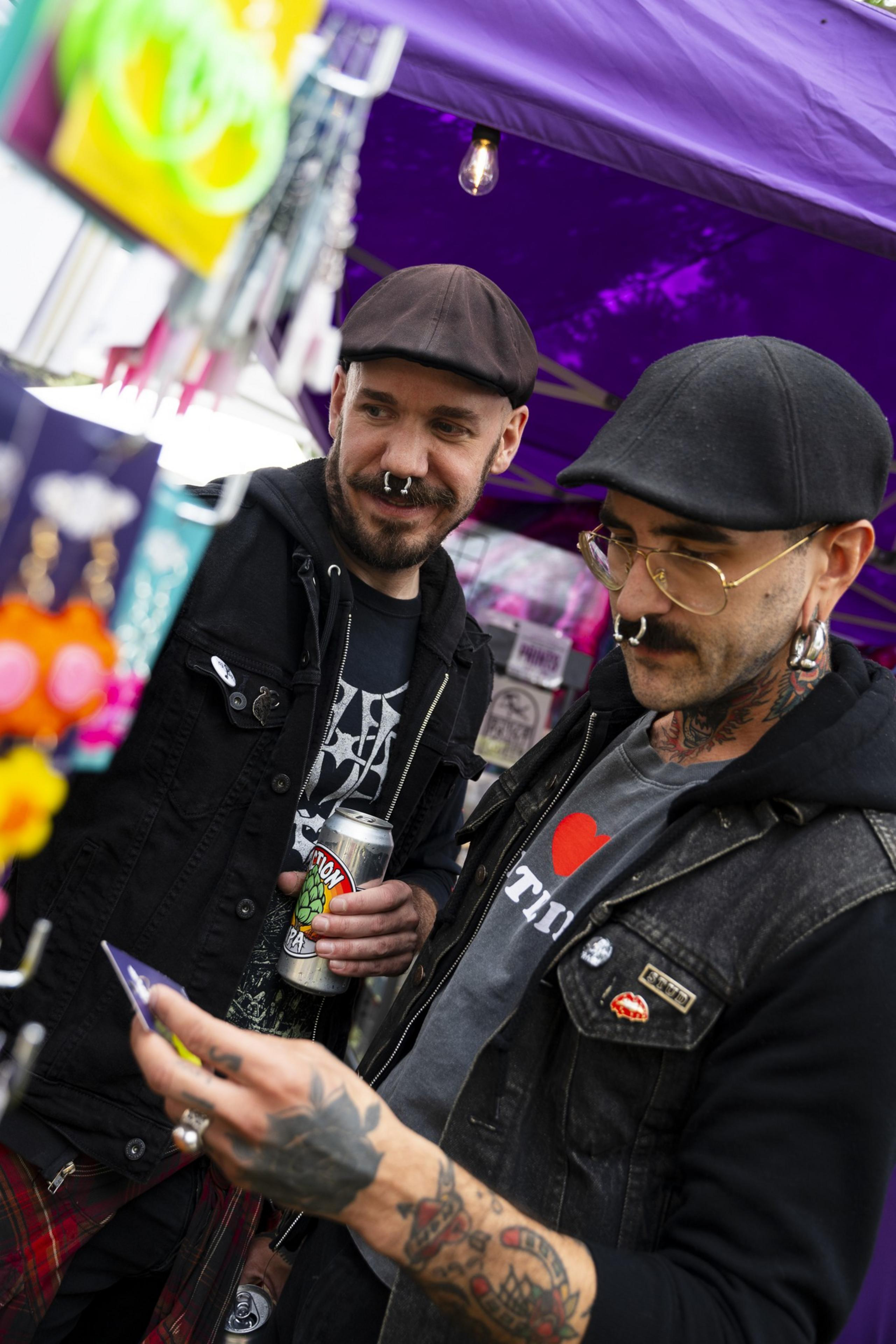 Two men with piercings and tattoos look at a display, one holding a can. They're under a purple tent, wearing hats and jackets with patches.