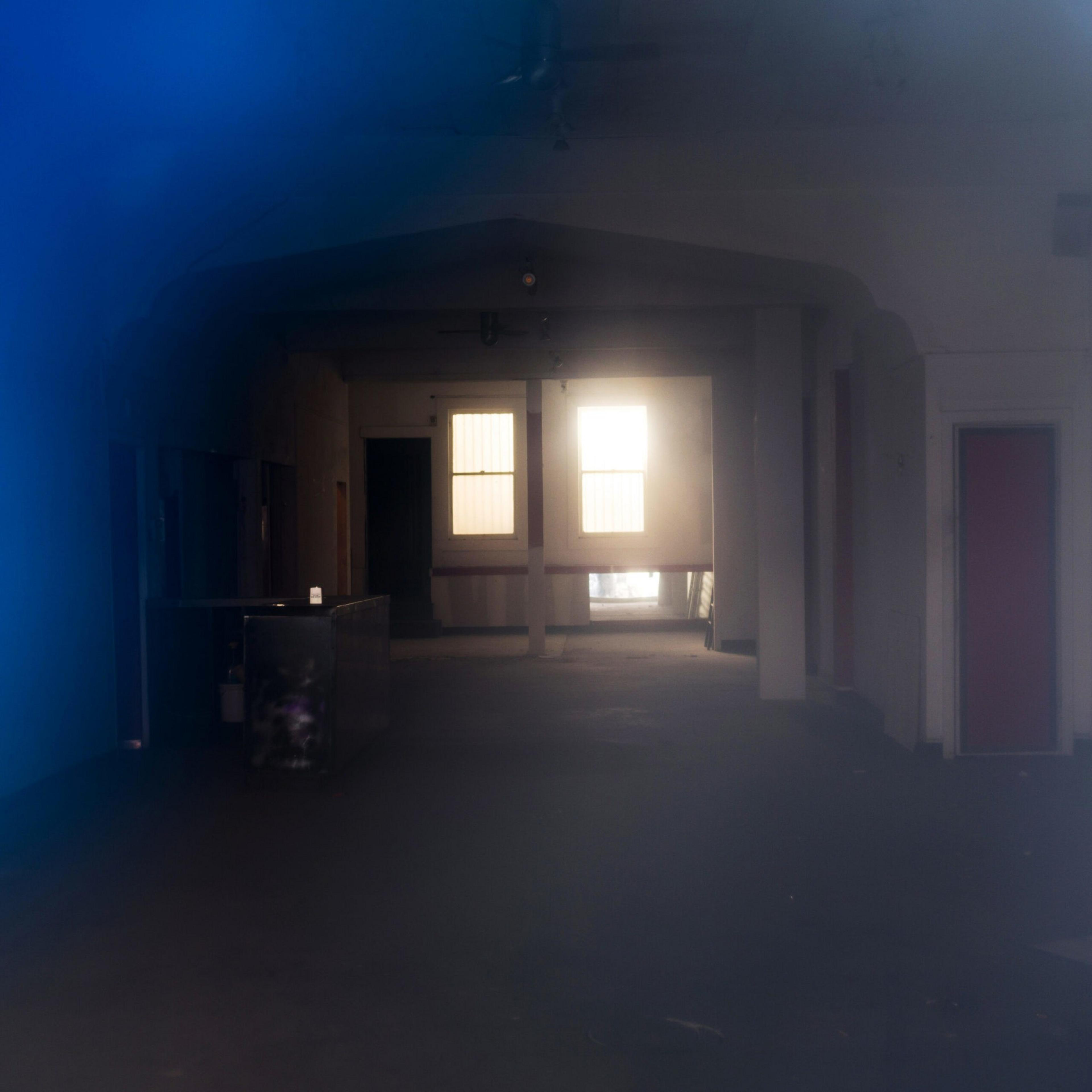 An empty, dimly lit room with two bright windows at the far end, a counter on the left, and several doorways along the walls.