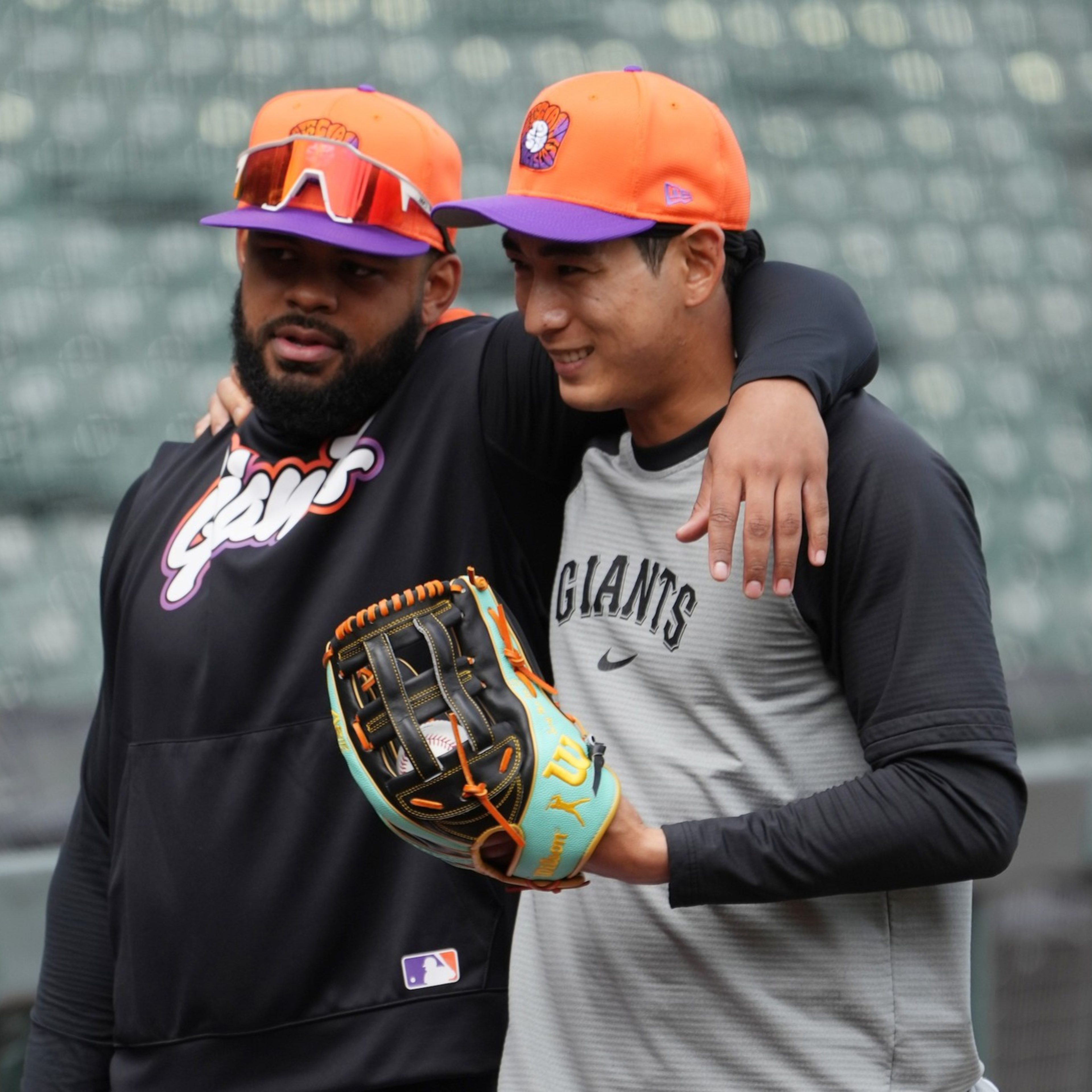 Two baseball players wearing orange caps with a purple logo share a friendly moment, one with his arm around the other’s shoulders.