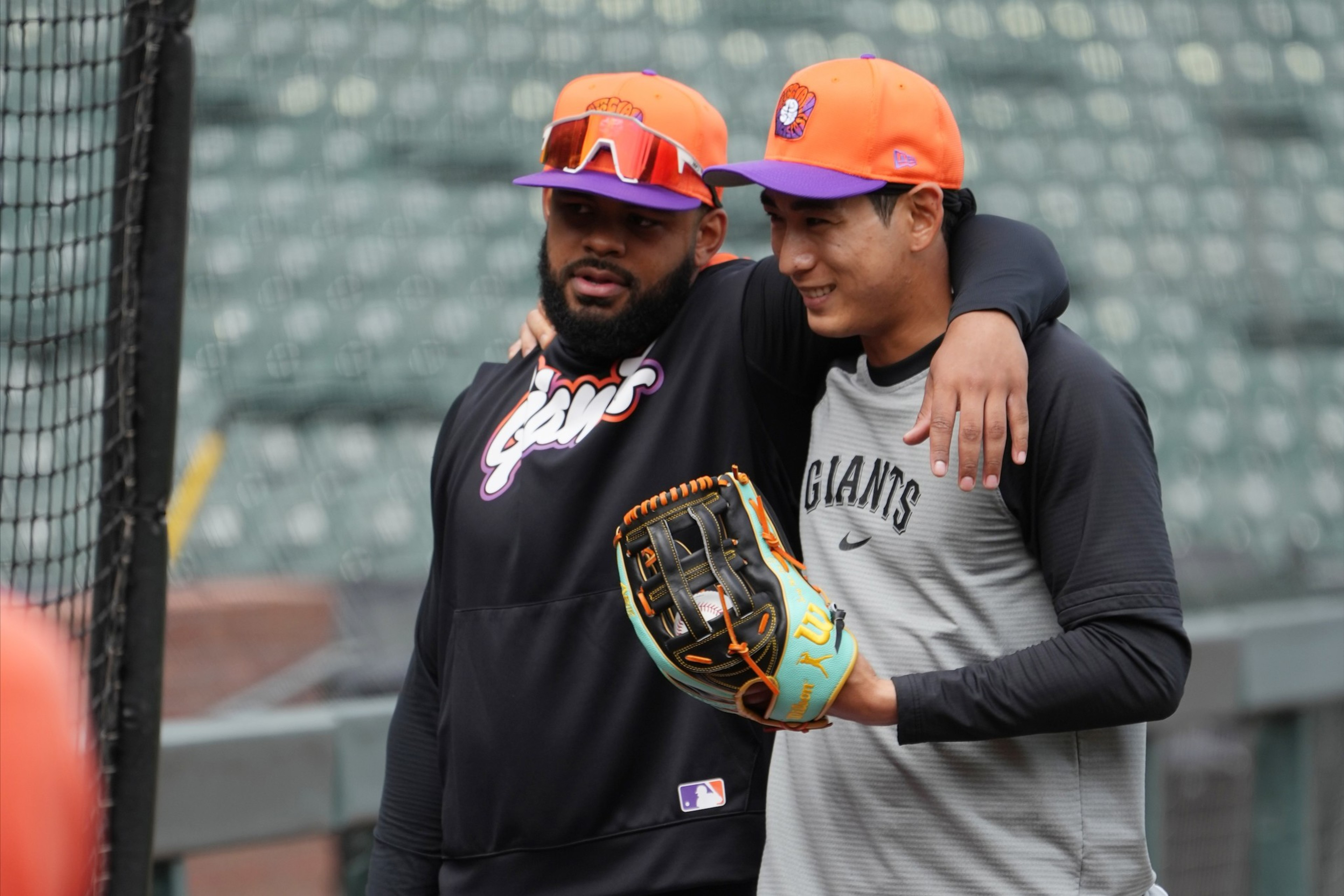 Two baseball players wearing orange caps with a purple logo share a friendly moment, one with his arm around the other’s shoulders.
