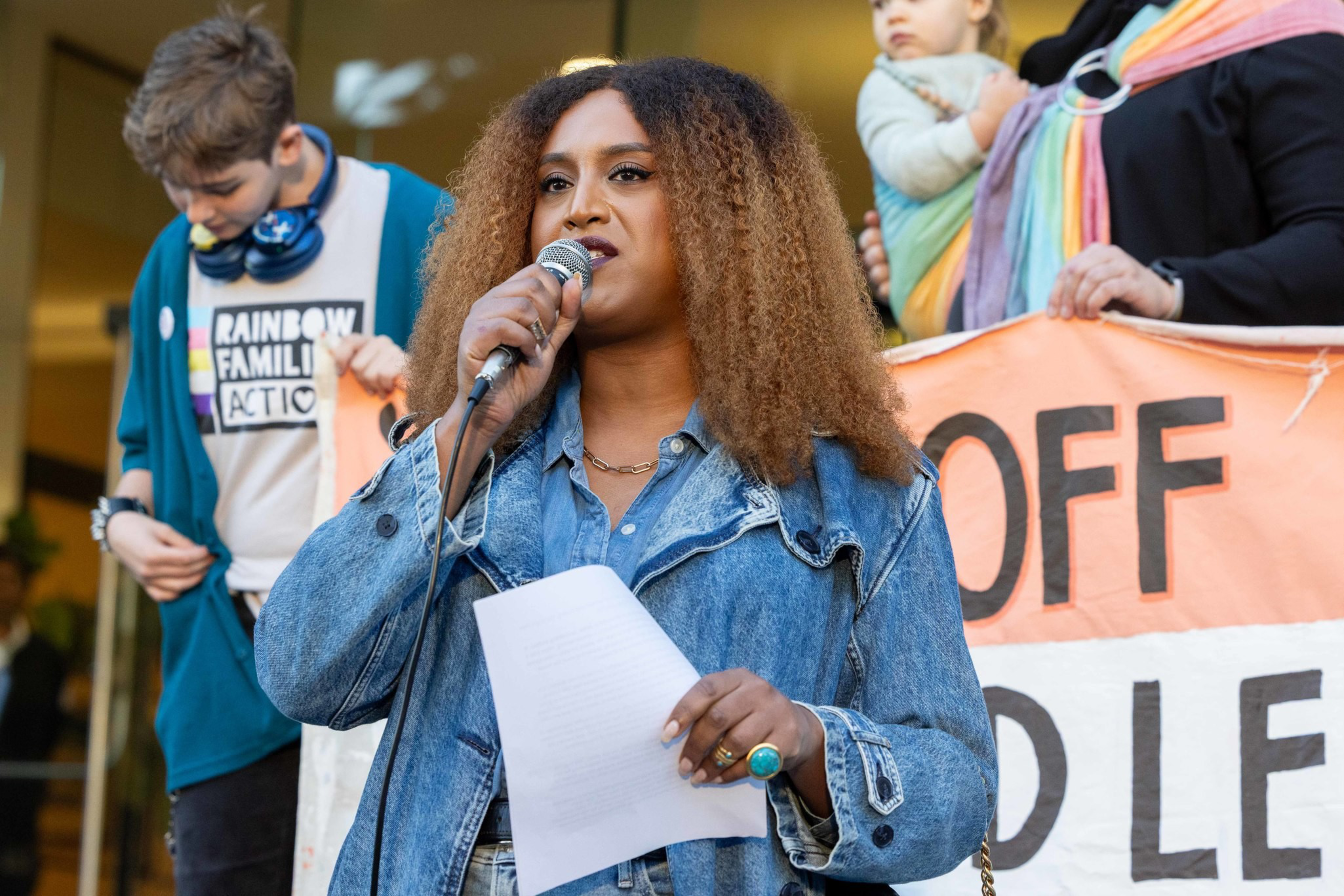 A woman with curly hair speaks into a microphone, holding a paper, while people behind hold a colorful banner and a child.
