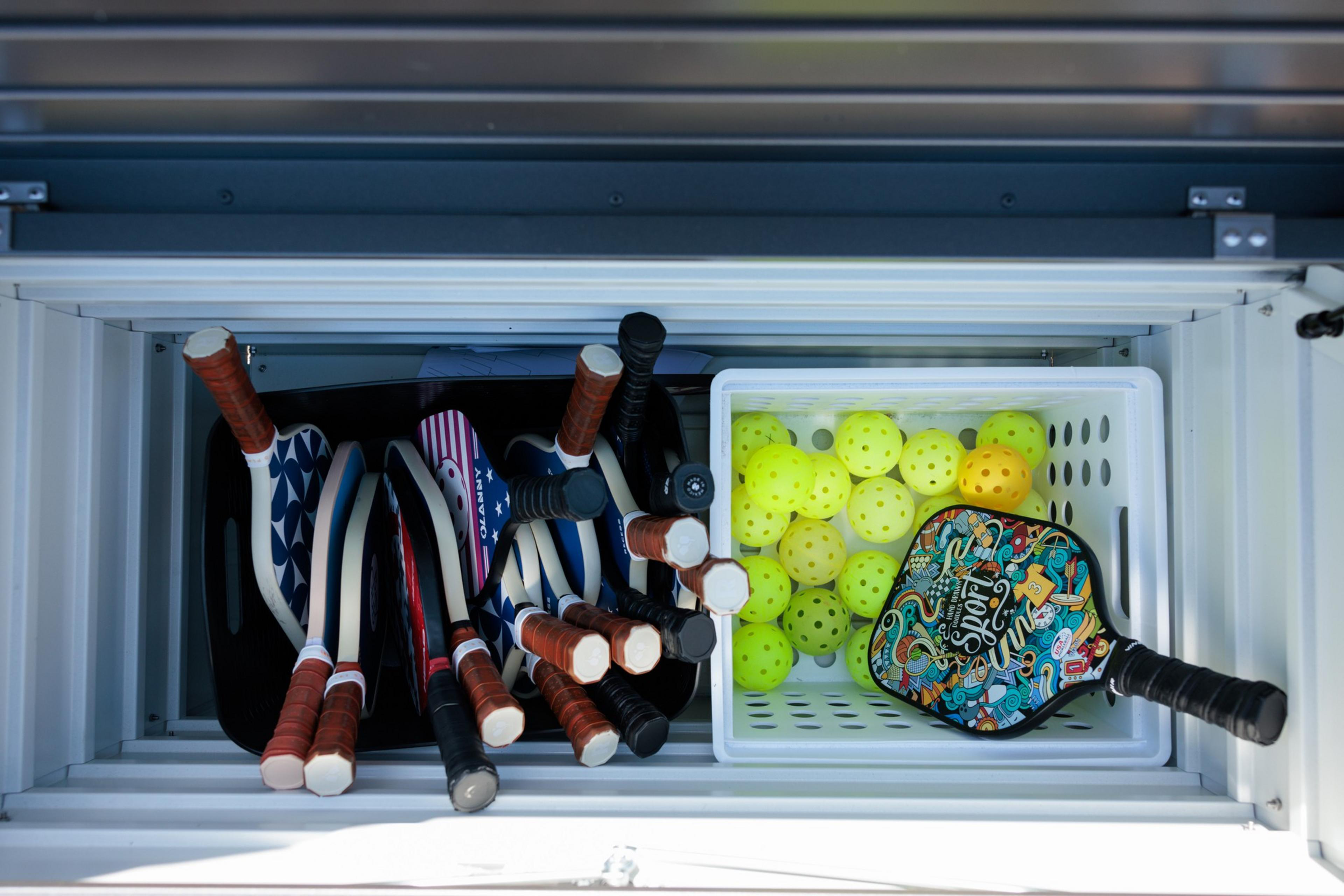 A bin holds pickleball paddles and a basket of yellow balls; one paddle has a colorful design.