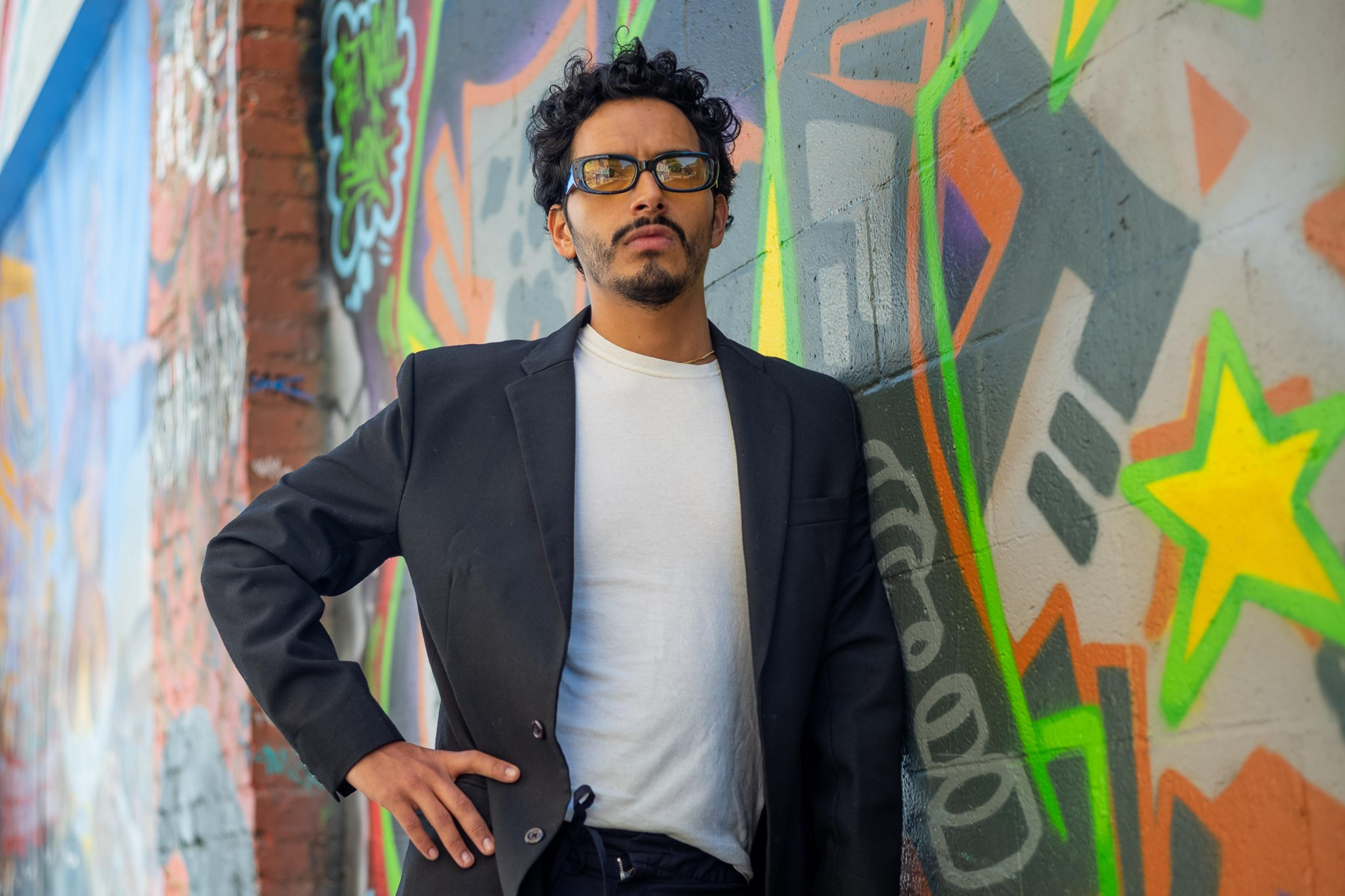 A man in a black blazer and white shirt stands confidently in front of a colorful graffiti-covered wall, with vibrant shapes and stars in the background.