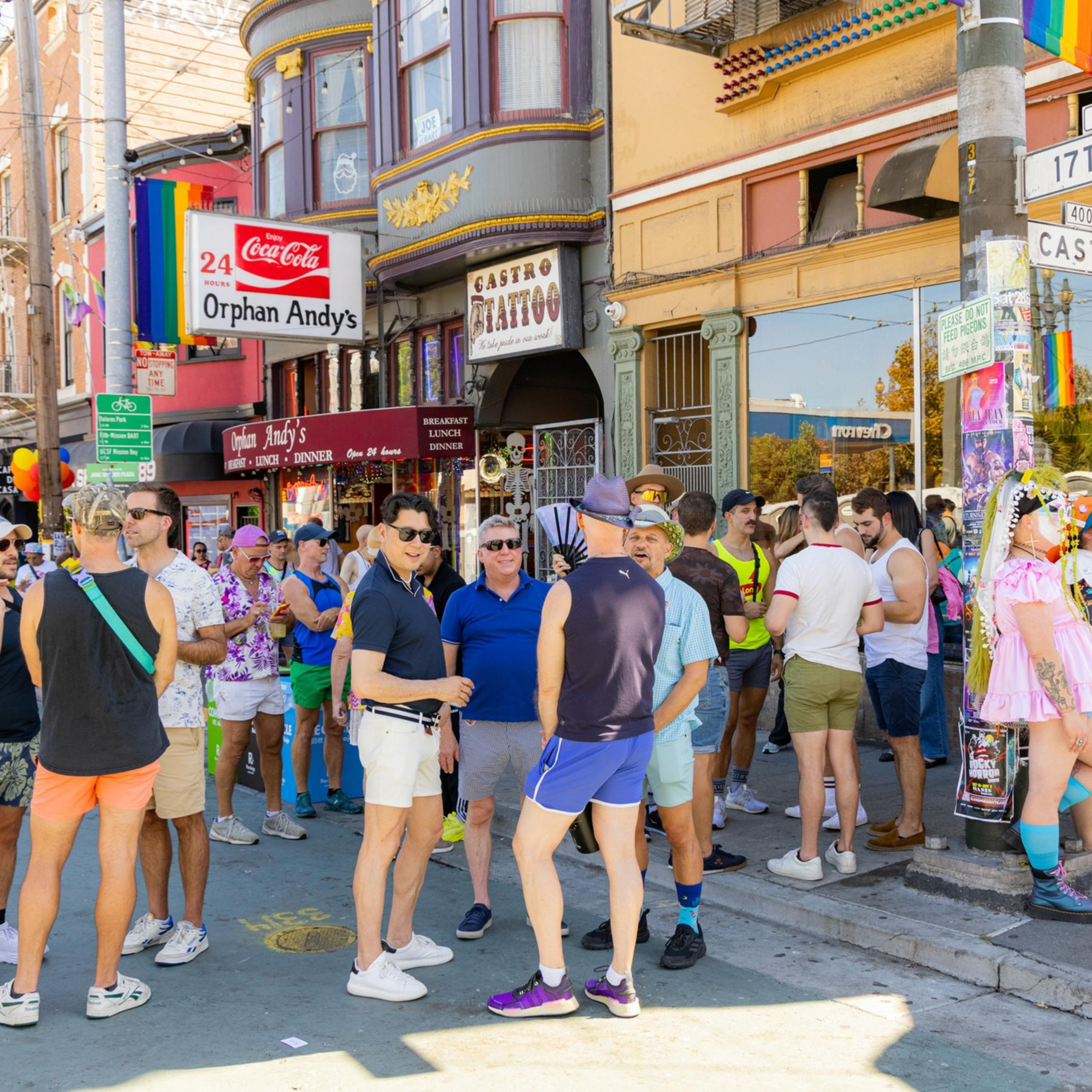 A lively group of people socializing on a colorful street corner with rainbow flags, vibrant outfits, and the sign for Castro Tattoo visible.
