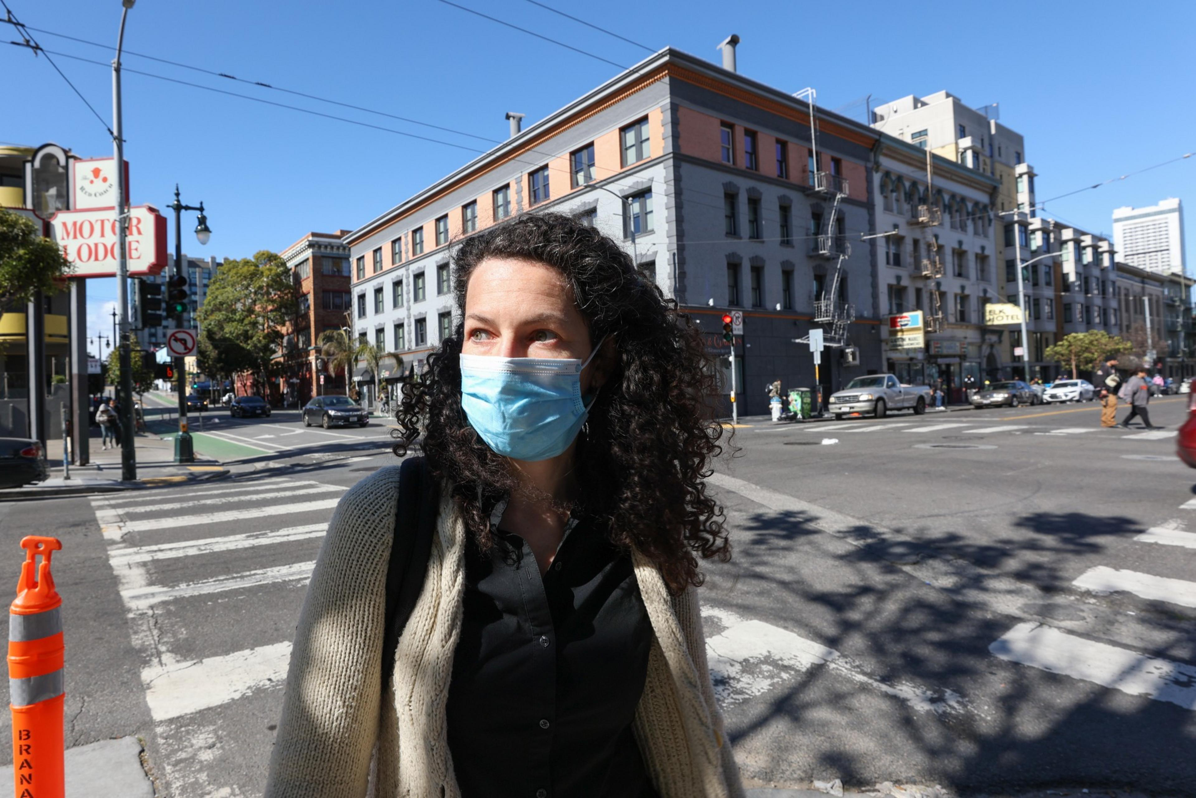 A woman with curly hair wearing a blue face mask, a beige cardigan, and a black shirt stands at a crosswalk in a sunny urban area.