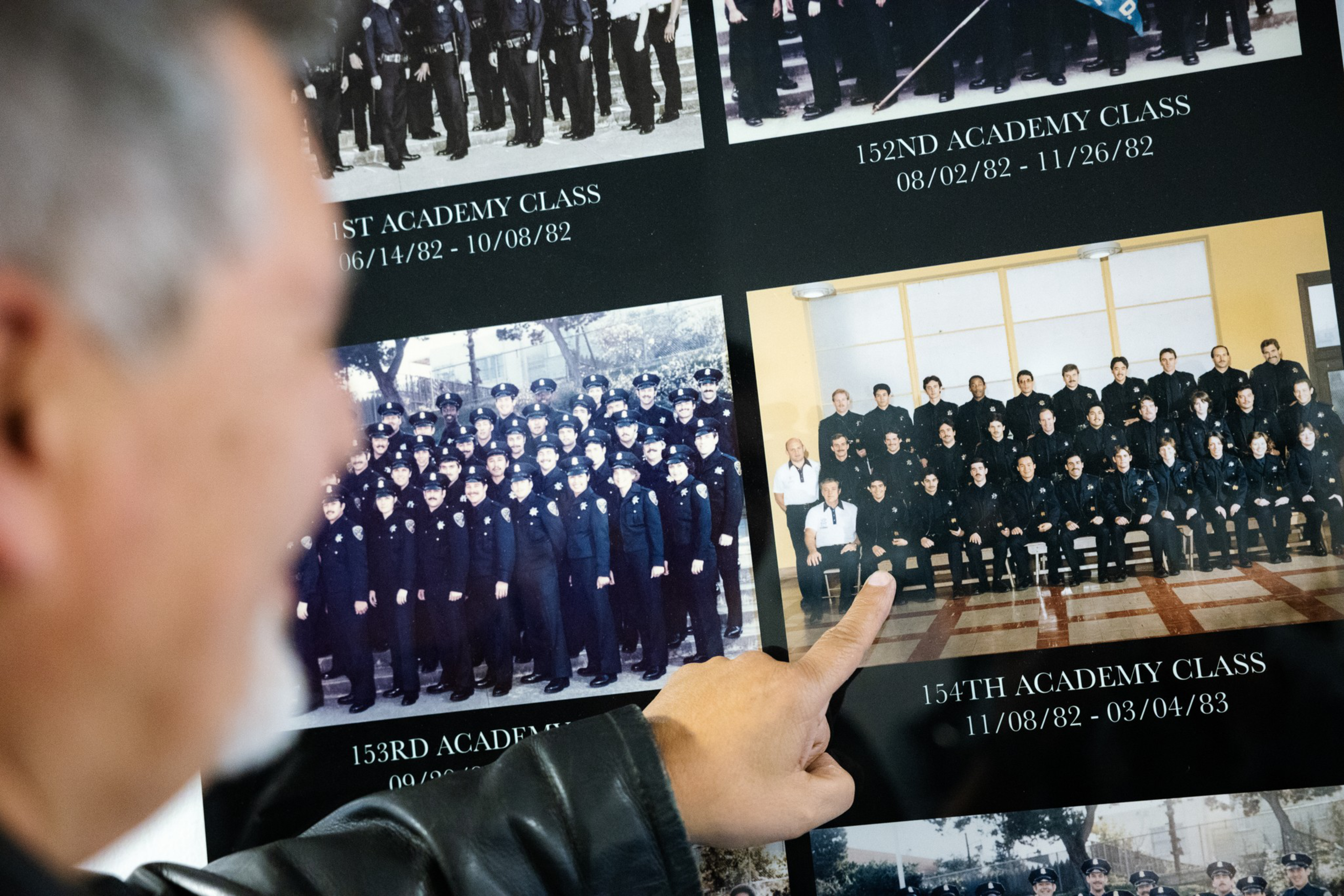 A person points to a photo of the 154th Academy Class, dated 11/08/82 - 03/04/83, among several class group photos on a display board.